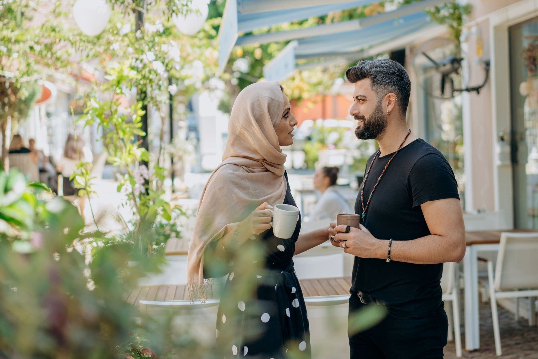 Uma mulher de lenço e um homem com barba conversam com chávenas na mão.
