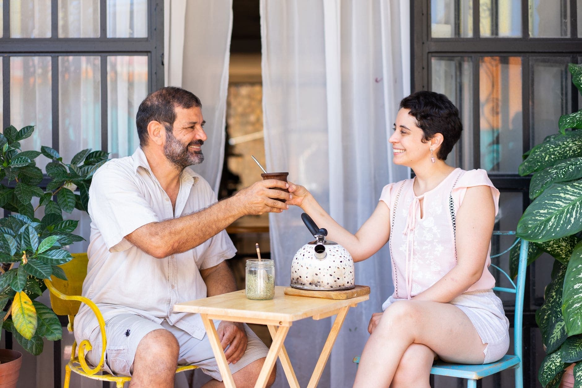 Um homem e uma mulher sorriem enquanto passam um chimarrão em uma varanda com plantas