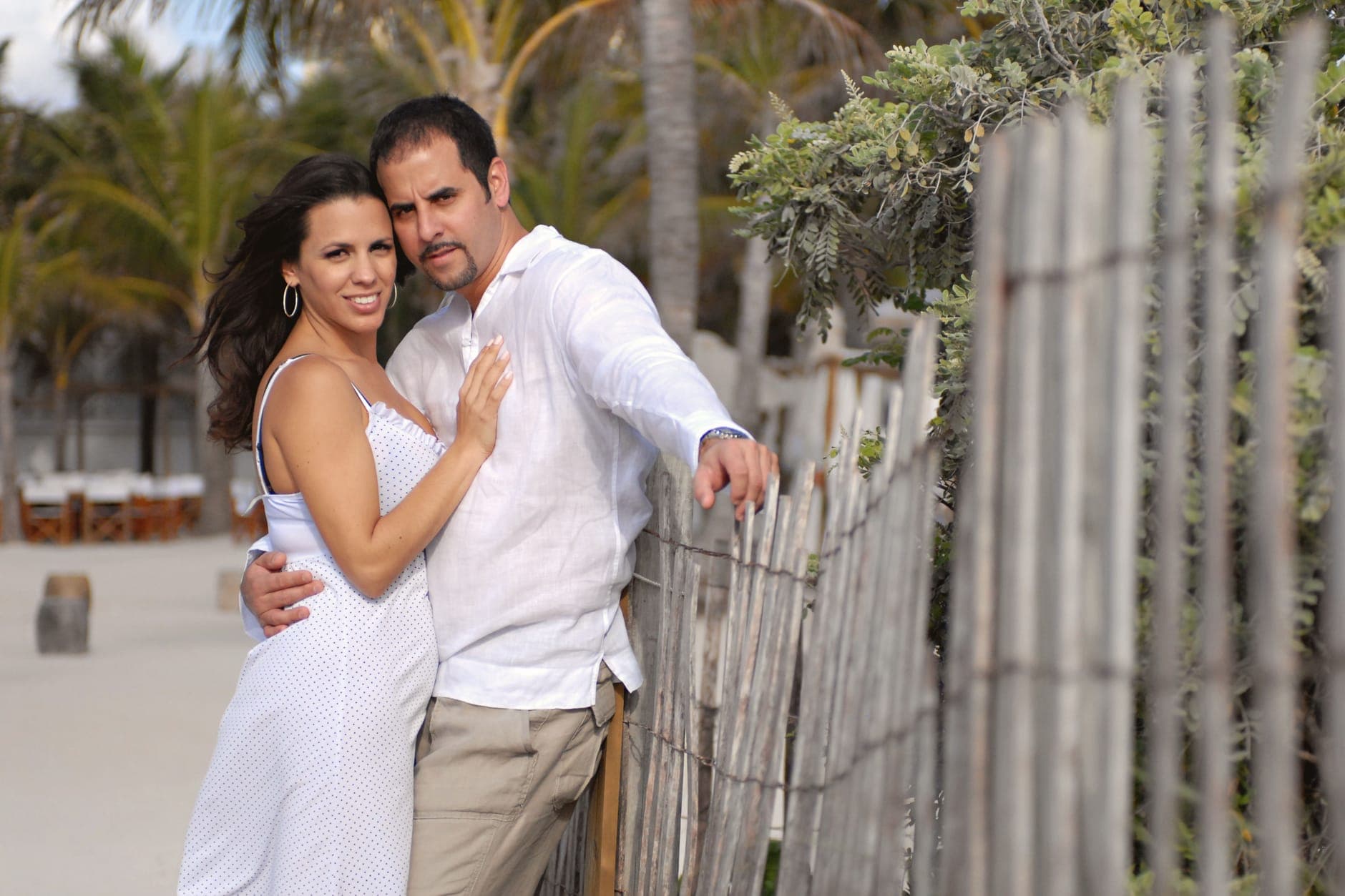 A man in a white linen shirt and a woman in a polka-dot sundress lean on a beach fence under palm trees.