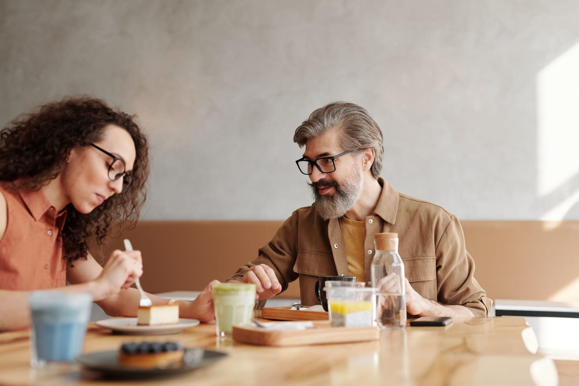 A gray-bearded man in glasses talks with a curly-haired woman over coffee and cake at a cafe table.