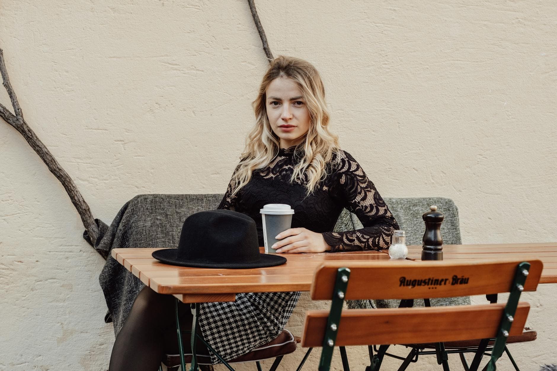 Blond kvinne i sort blondetopp sitter ved et kafébord med kaffekopp og sort hatt.