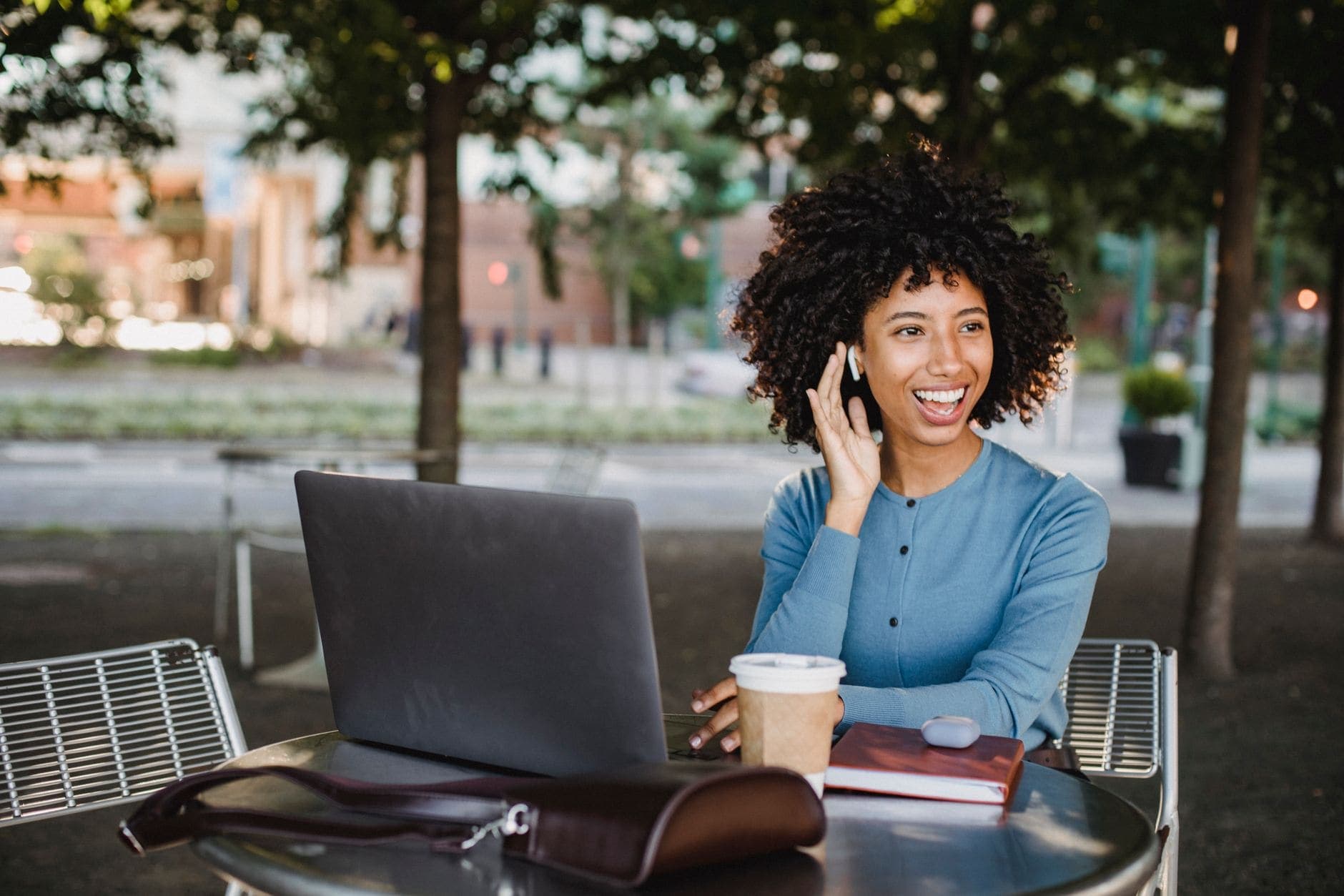 A smiling woman in blue holds a hand beside an earbud at an outdoor table with a laptop.