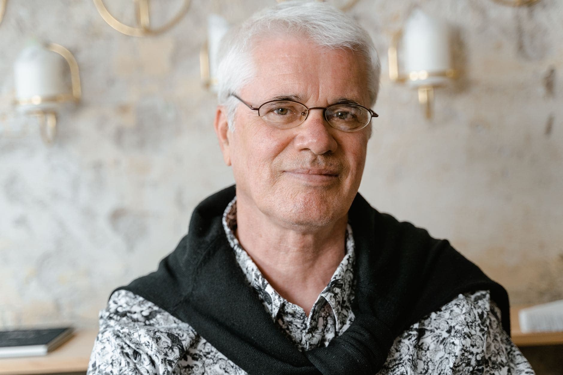 A man with glasses and white hair looks towards the camera in a patterned shirt.