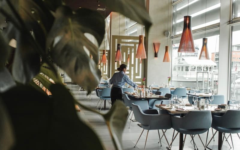 Lys restaurant med grå stole og pendellamper, en tjener dækker bord ved de store vinduer.