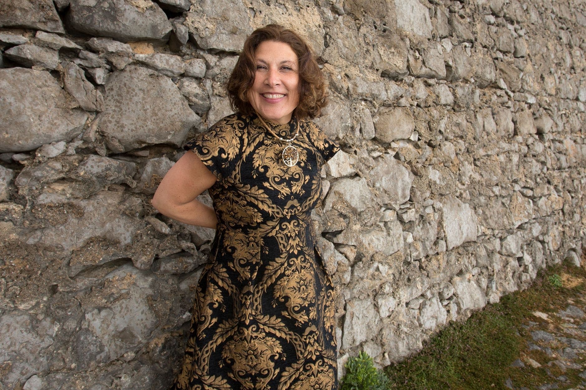 Uma mulher de vestido preto e dourado sorri junto a um muro de pedra.