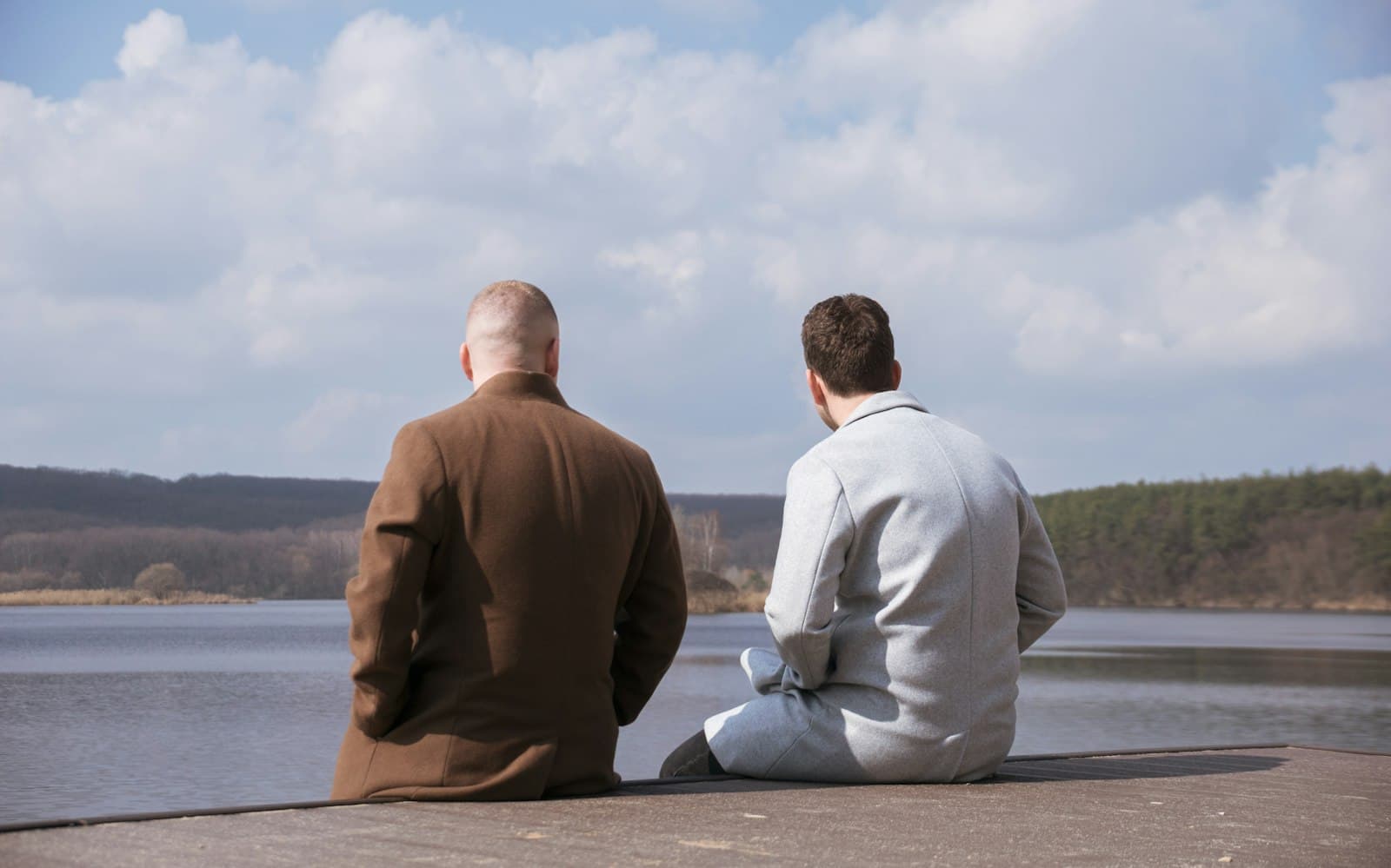 Zwei Männer in Mänteln sitzen von hinten gesehen auf einem Steg und blicken über einen See