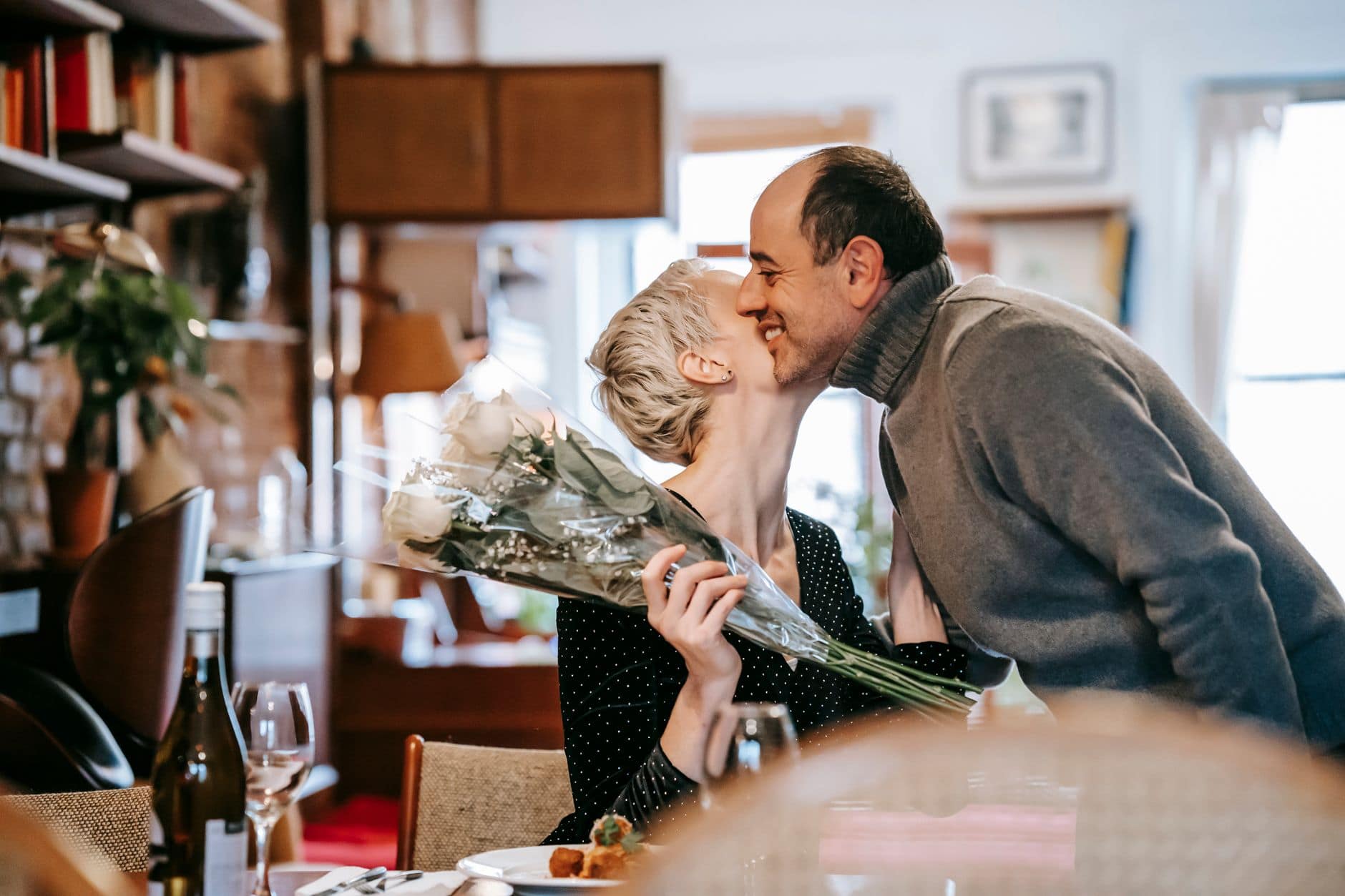 Eine Frau mit weißen Rosen küsst einen lächelnden Mann im Restaurant auf die Wange.