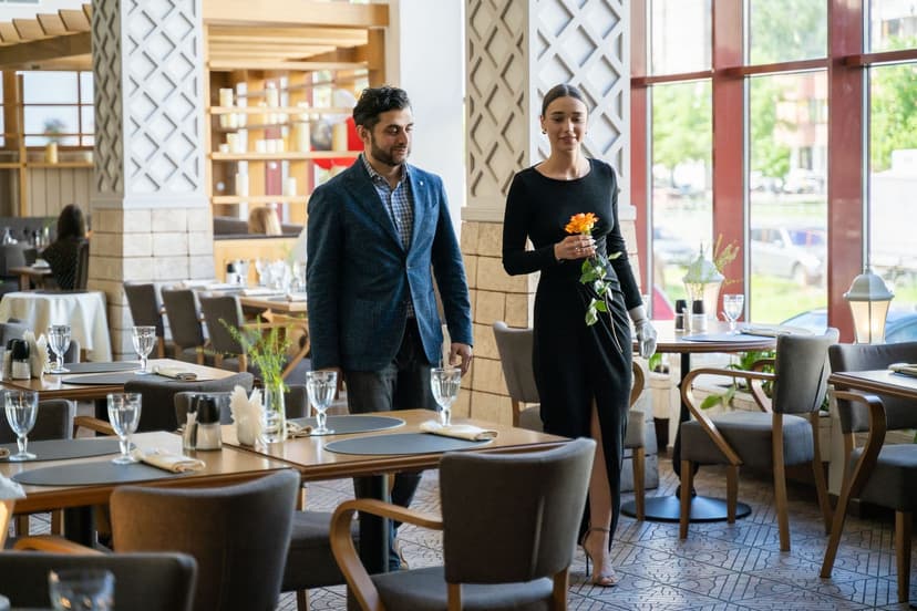 Velklædt par på vej gennem en stilfuld restaurant - manden i blå blazer og ternet skjorte, kvinden i lang sort kjole med en enkelt orange rose i hånden