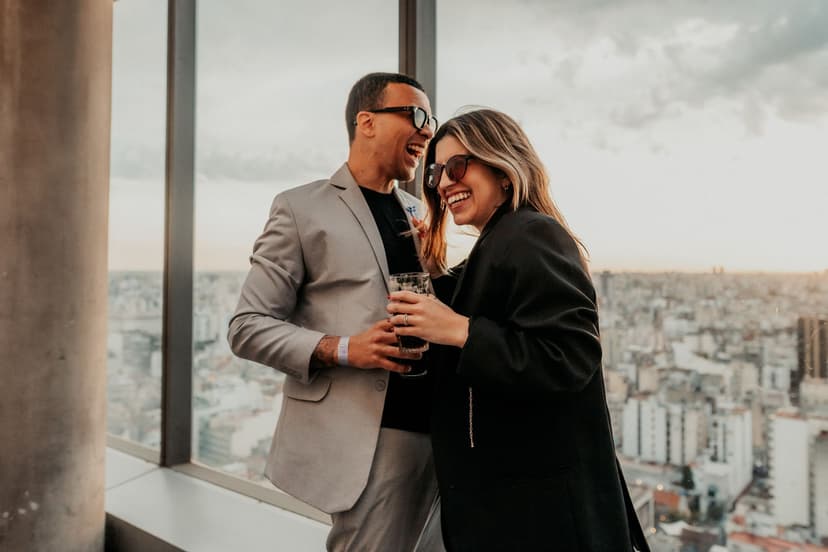 Grinende, velklædt par i lys og mørk blazer skåler i drinks på en rooftop-bar med panoramaudsigt over byens tage i det gyldne aftenlys