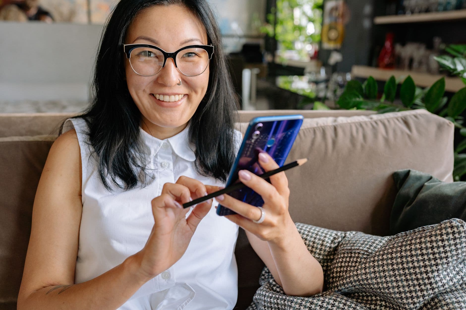 Eine lächelnde Frau mit Brille sitzt auf einem Sofa und hält ihr Smartphone und einen Stift.