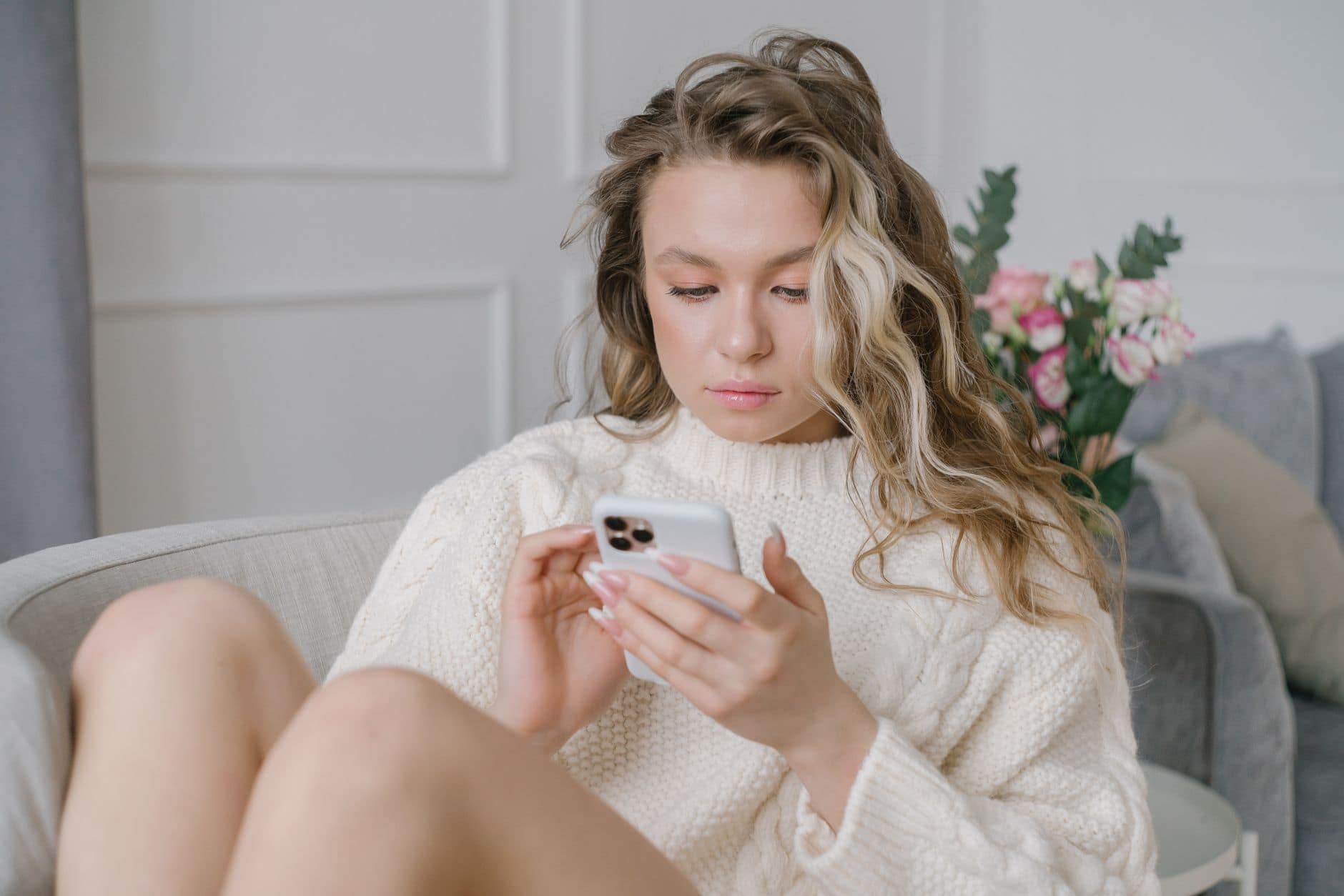 Eine Frau im hellen Strickpullover sitzt auf einem Sofa und schaut auf ihr Smartphone.