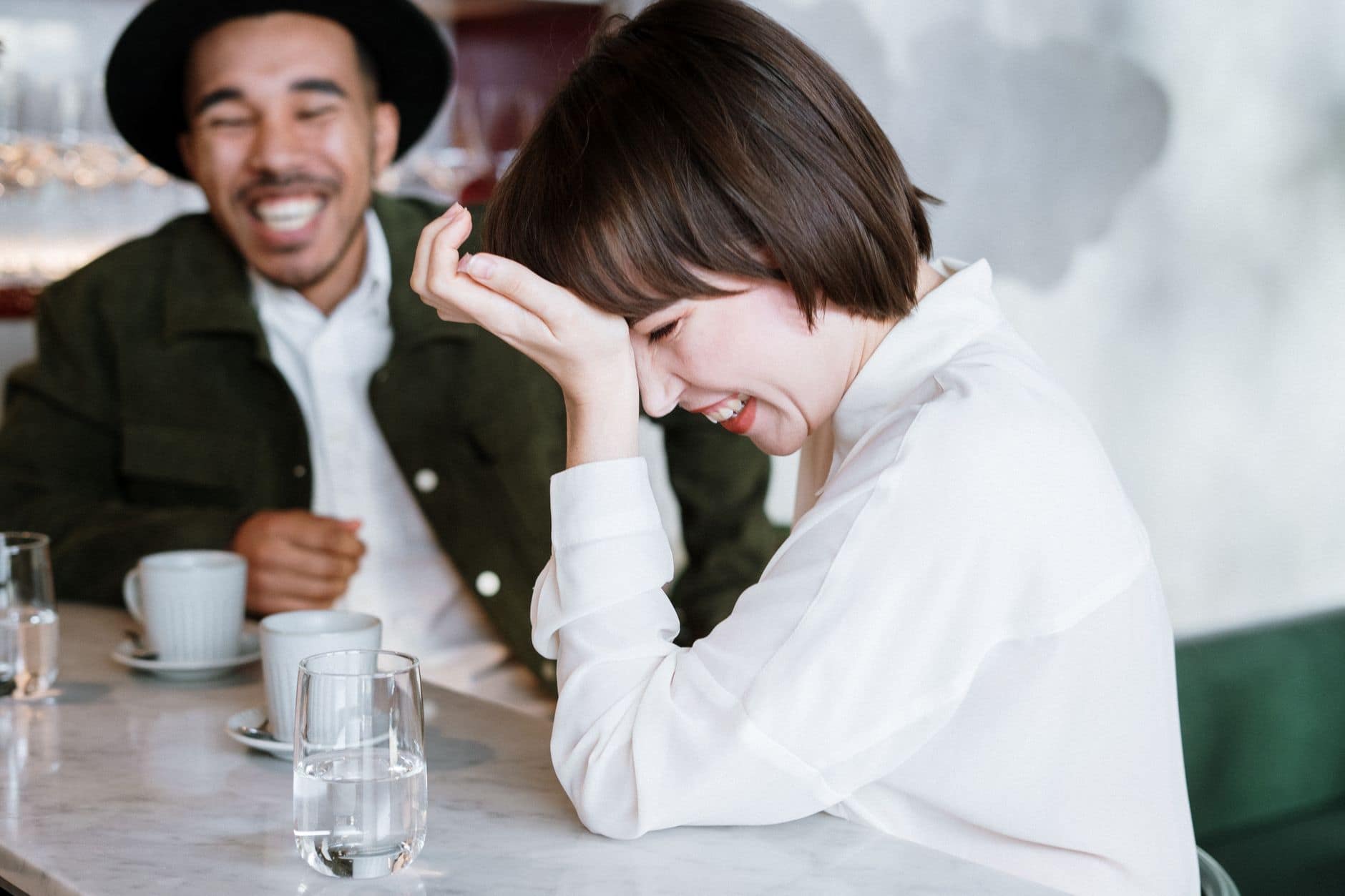 Ein Mann mit schwarzem Hut und eine Frau in weisser Bluse lachen gemeinsam im Café.