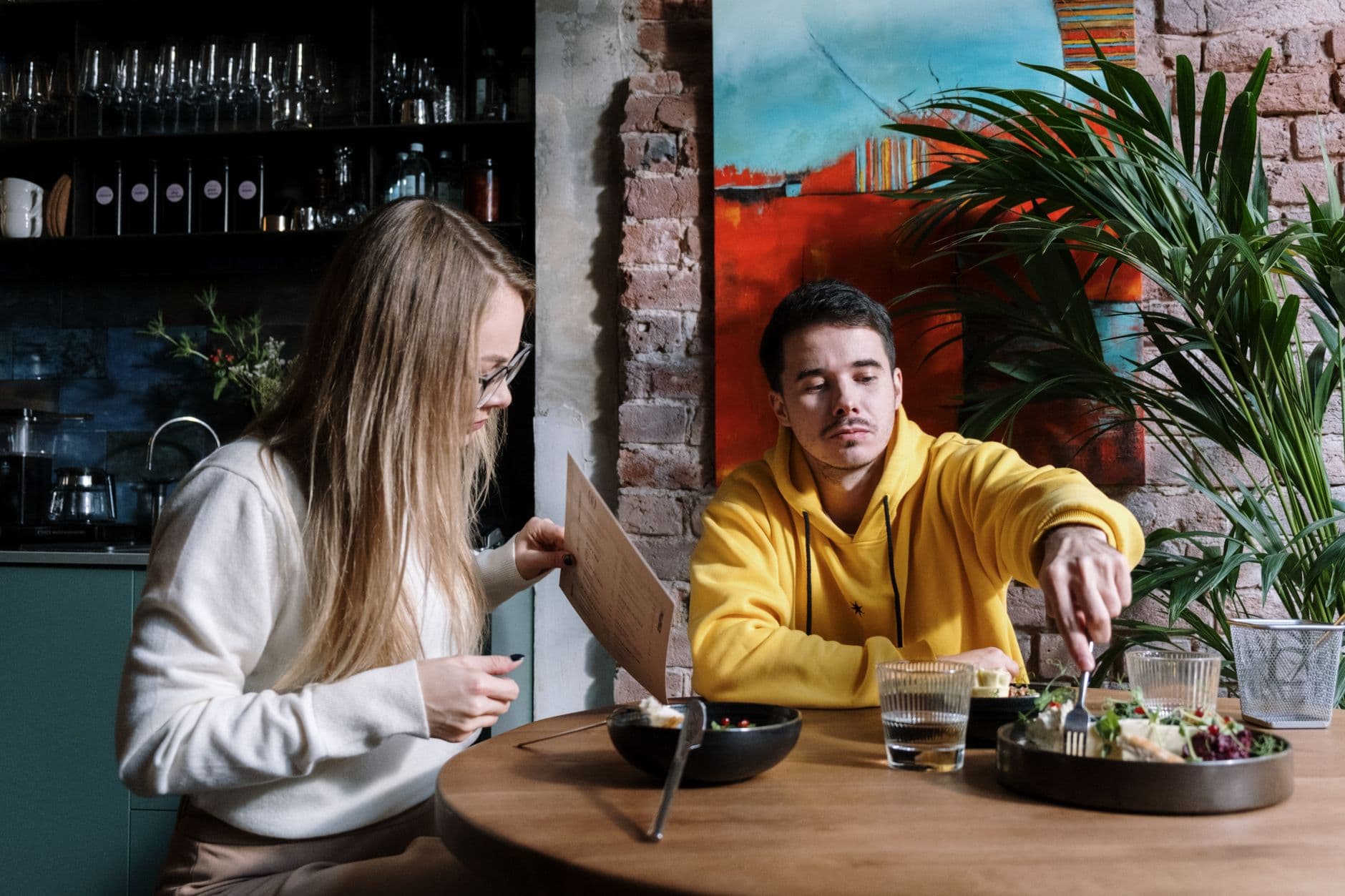 Een man in een felgele hoodie en een vrouw met een bril zitten samen te eten aan een tafeltje in een restaurant.