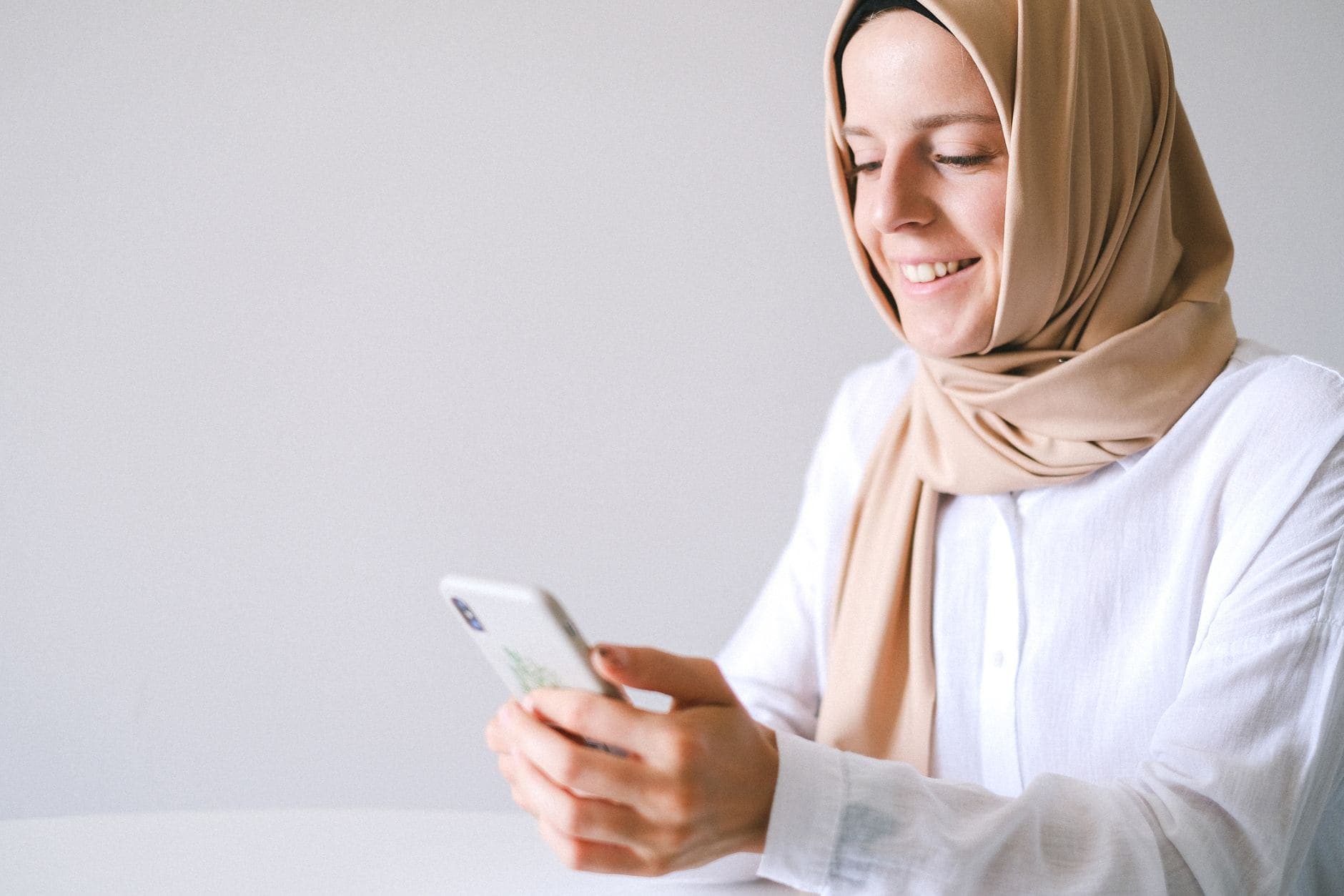 A woman in a beige headscarf smiles while looking at her phone.