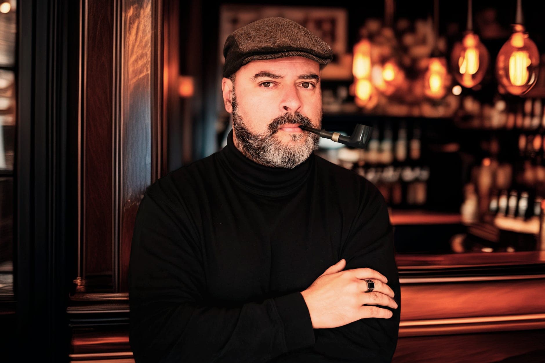 A bearded man in a flat cap holds a pipe in his mouth, arms folded, in a warm-lit bar.