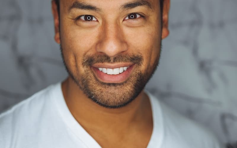 Close-up of a smiling man with a short beard in a white V-neck t-shirt against a grey wall.