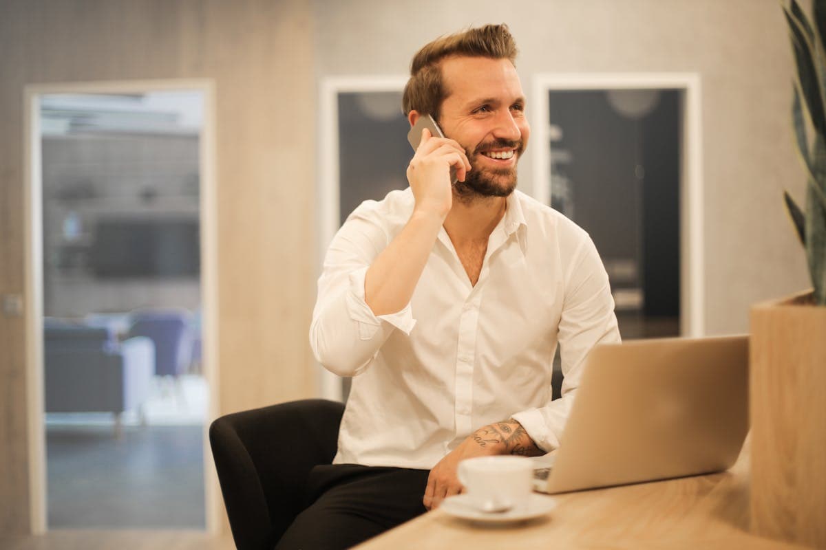 Smilende mand i hvid skjorte taler i telefon ved et skrivebord med bærbar og kaffekop.