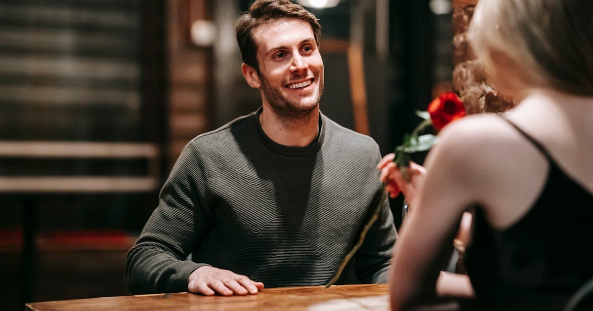 Smilende mand i mørk sweatshirt sidder over for en person med rød rose ved et træbord.