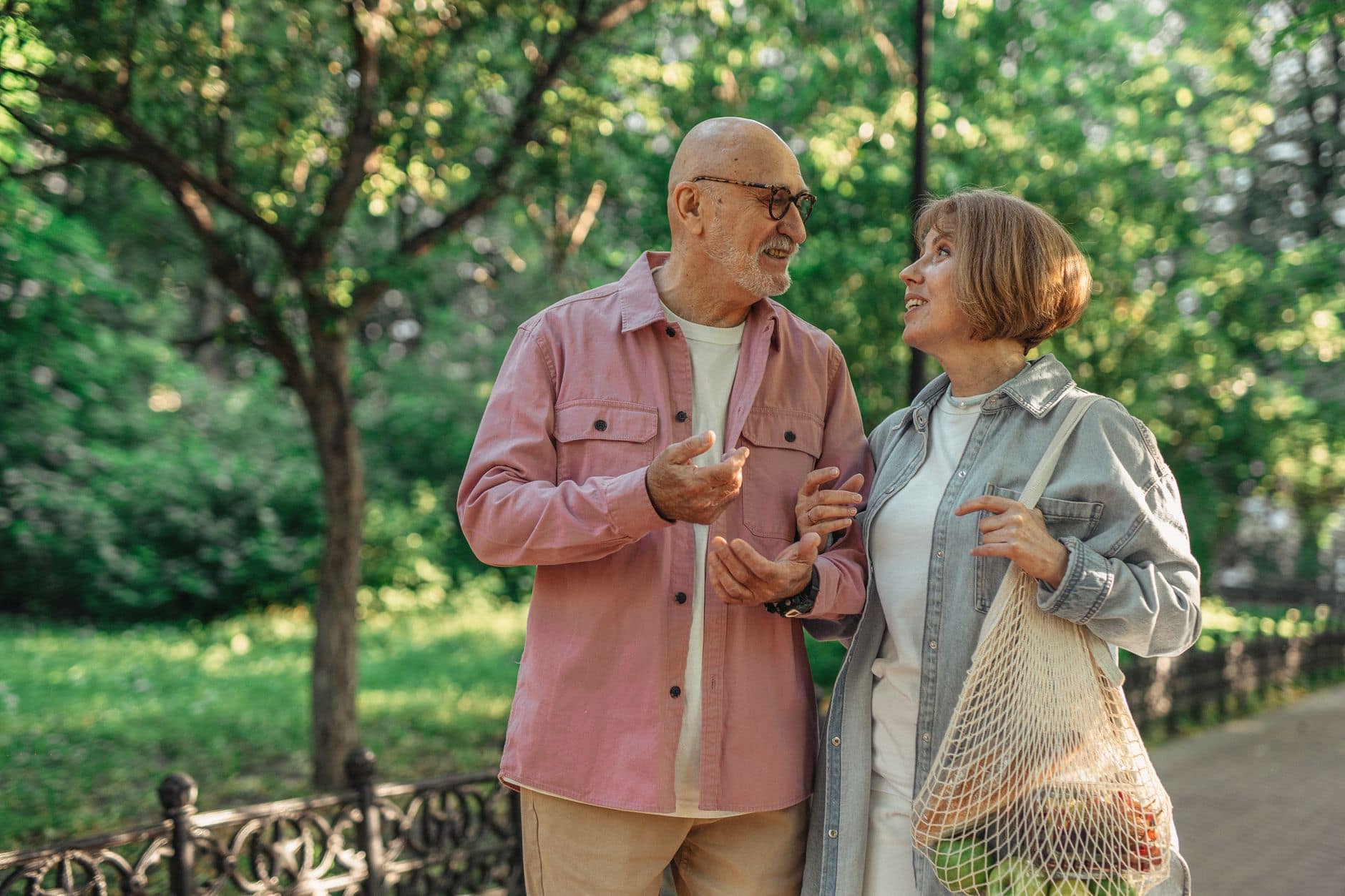 Casal de idosos caminha e conversa em um parque arborizado em um dia de sol.