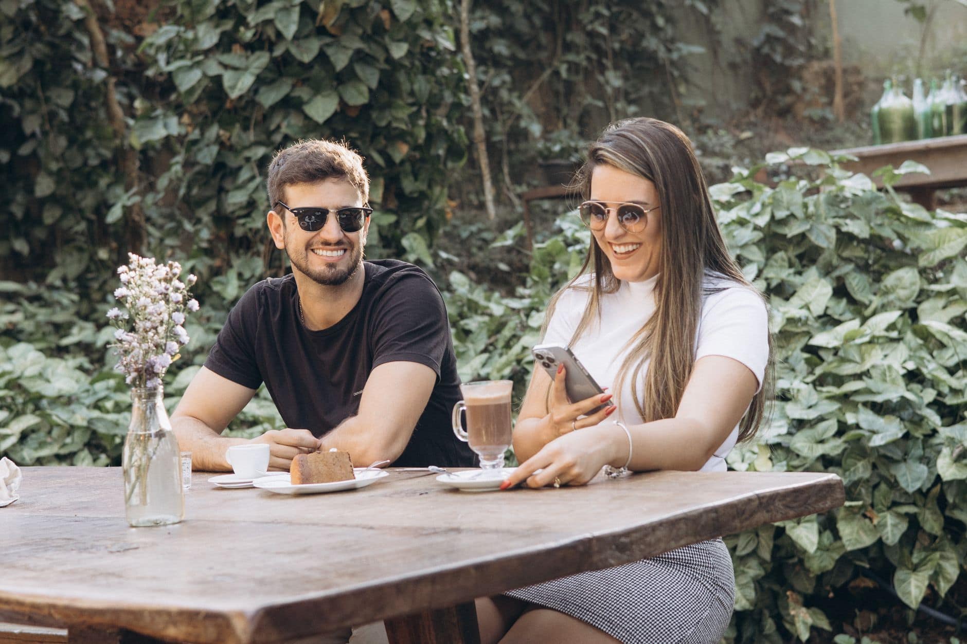 Una coppia sorride a un tavolo all'aperto con caffè e dolce; la donna tiene un telefono.