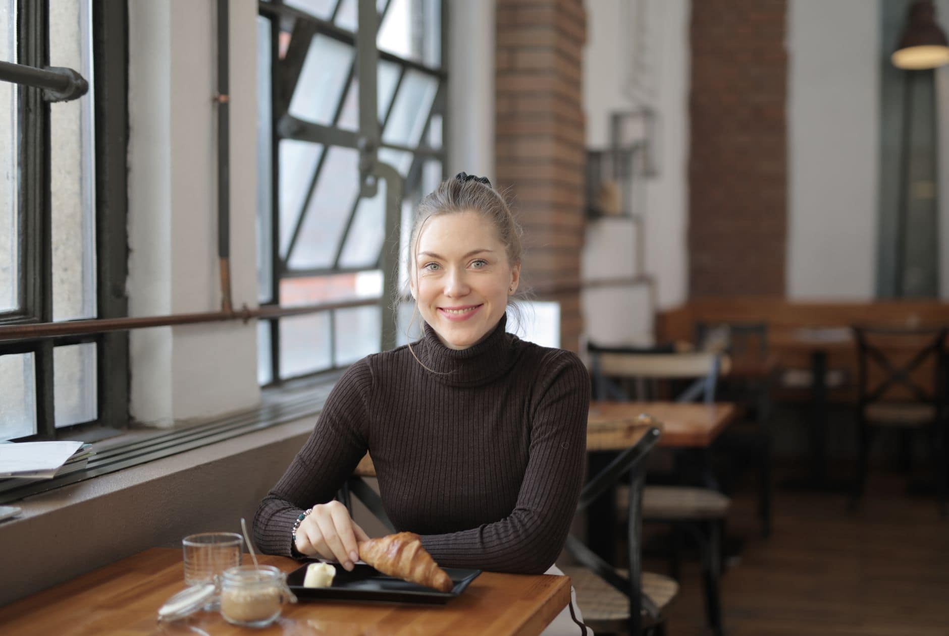 Una donna con un dolcevita scuro sorride al tavolo di un bar vicino alla finestra.