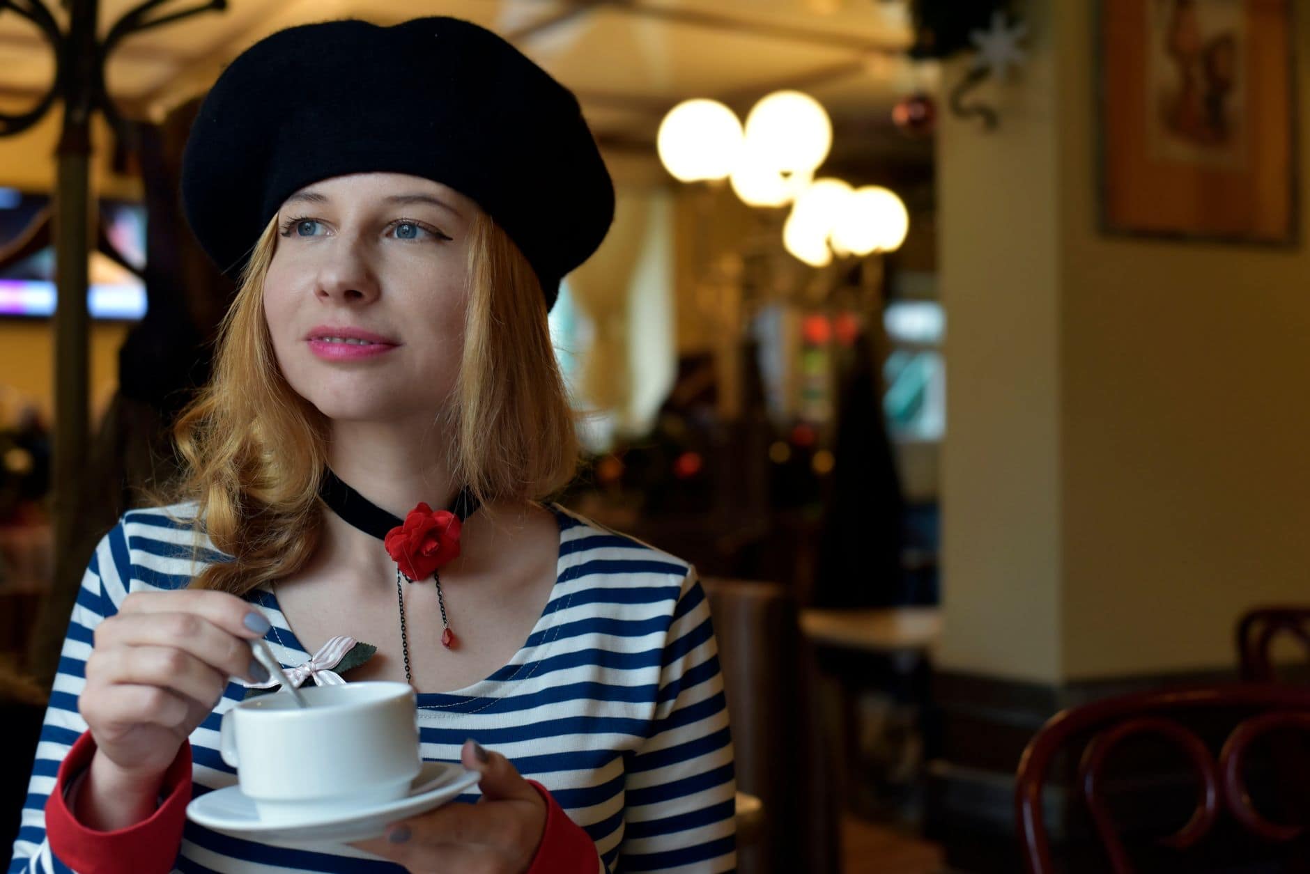 O femeie blondă cu beretă neagră și bluză în dungi ține o ceașcă și o farfurioară în cafenea