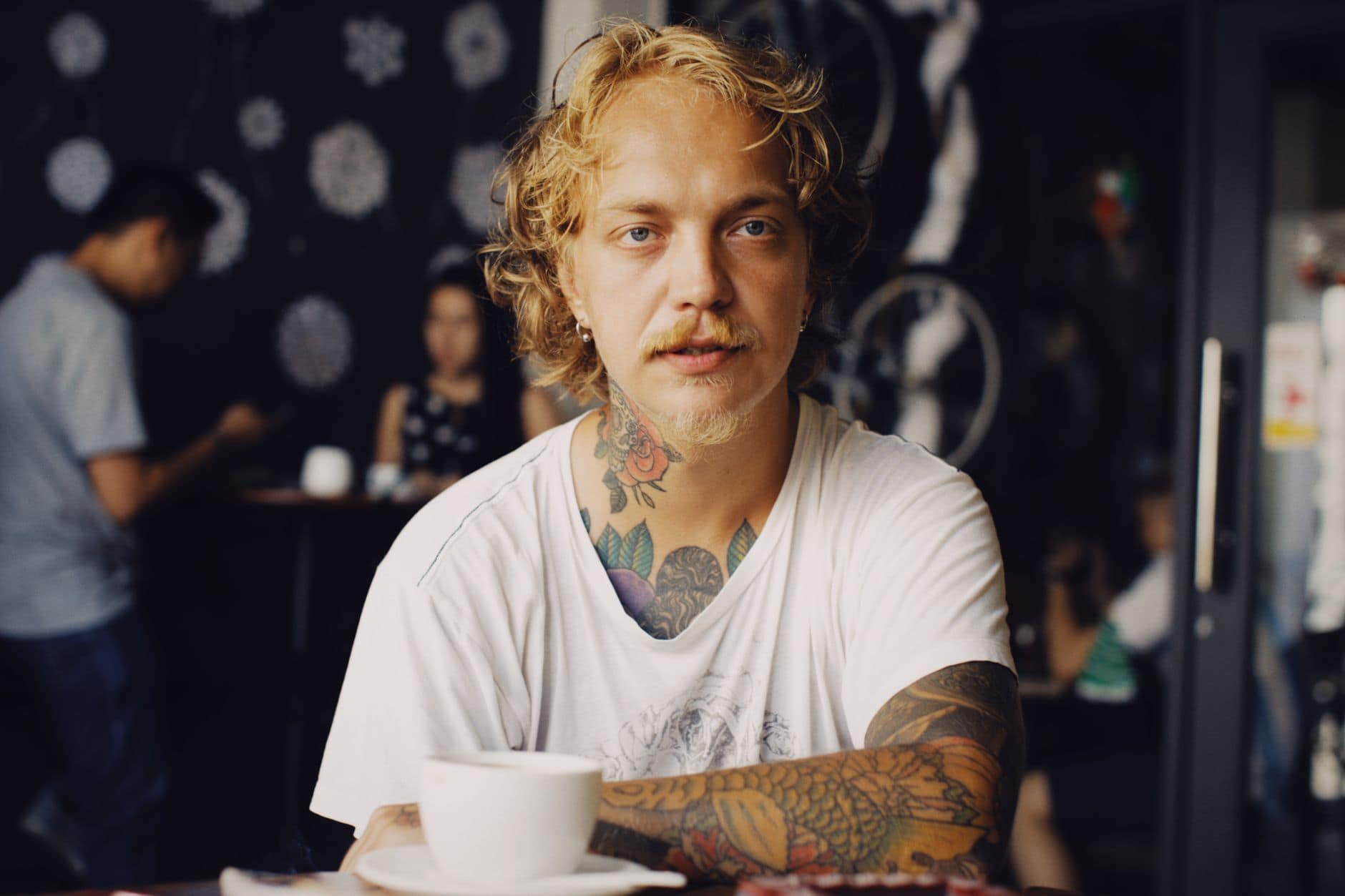 Un bărbat blond cu mustață și tatuaje stă la o masă cu o ceașcă albă de cafea
