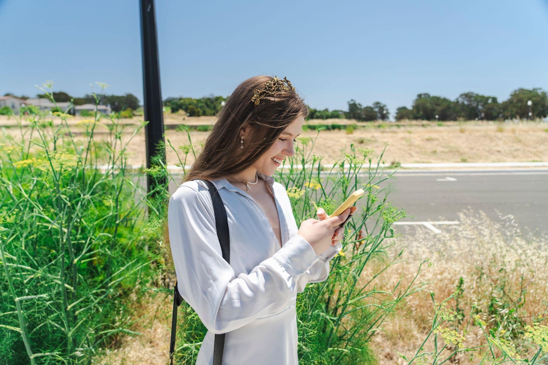 O femeie zâmbitoare în cămașă deschisă la culoare privește telefonul lângă plante înalte