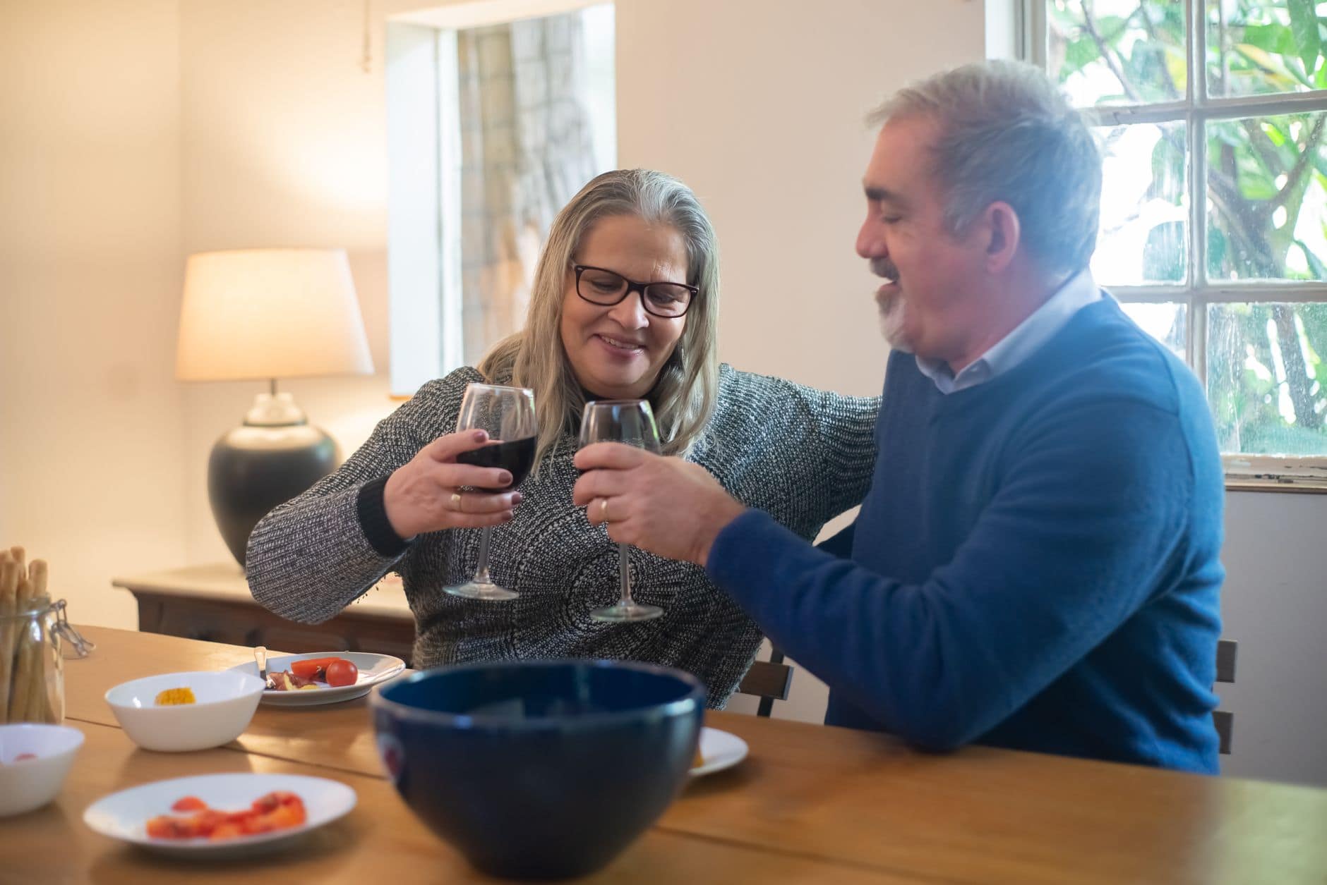 Dos personas adultas sonríen mientras brindan con vino junto a una ventana.