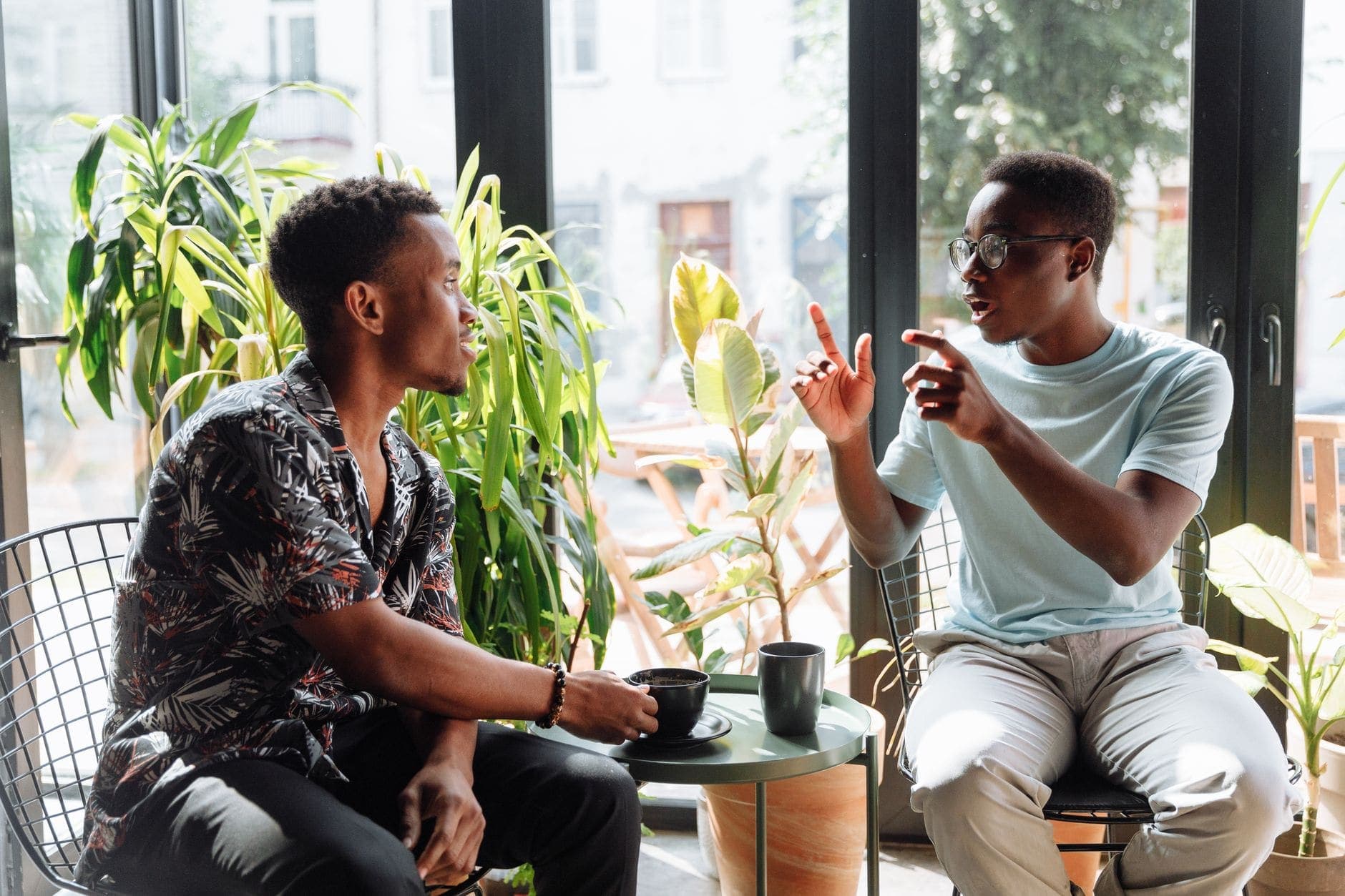 Dois homens conversam junto a uma janela; um deles gesticula sobre uma mesa com duas chávenas.
