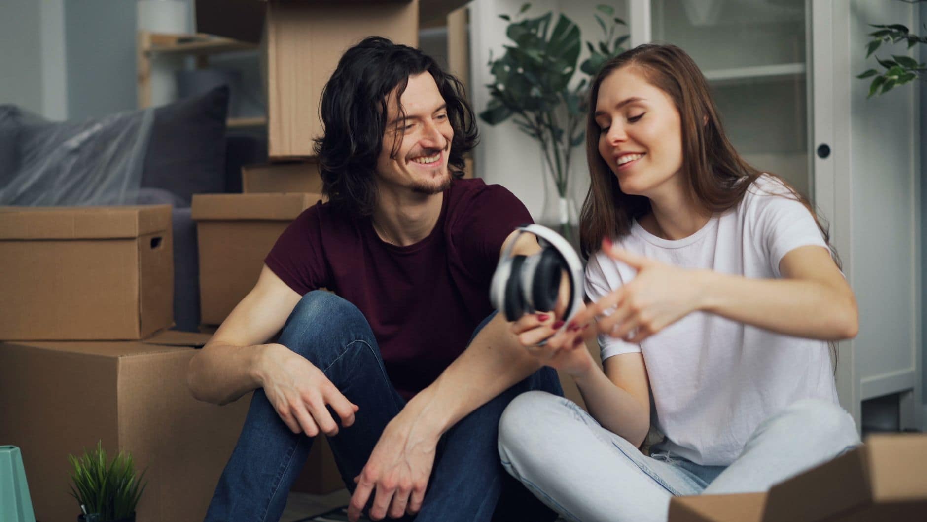 Casal jovem sorri sentado no chão entre caixas de mudança, segurando fones de ouvido.