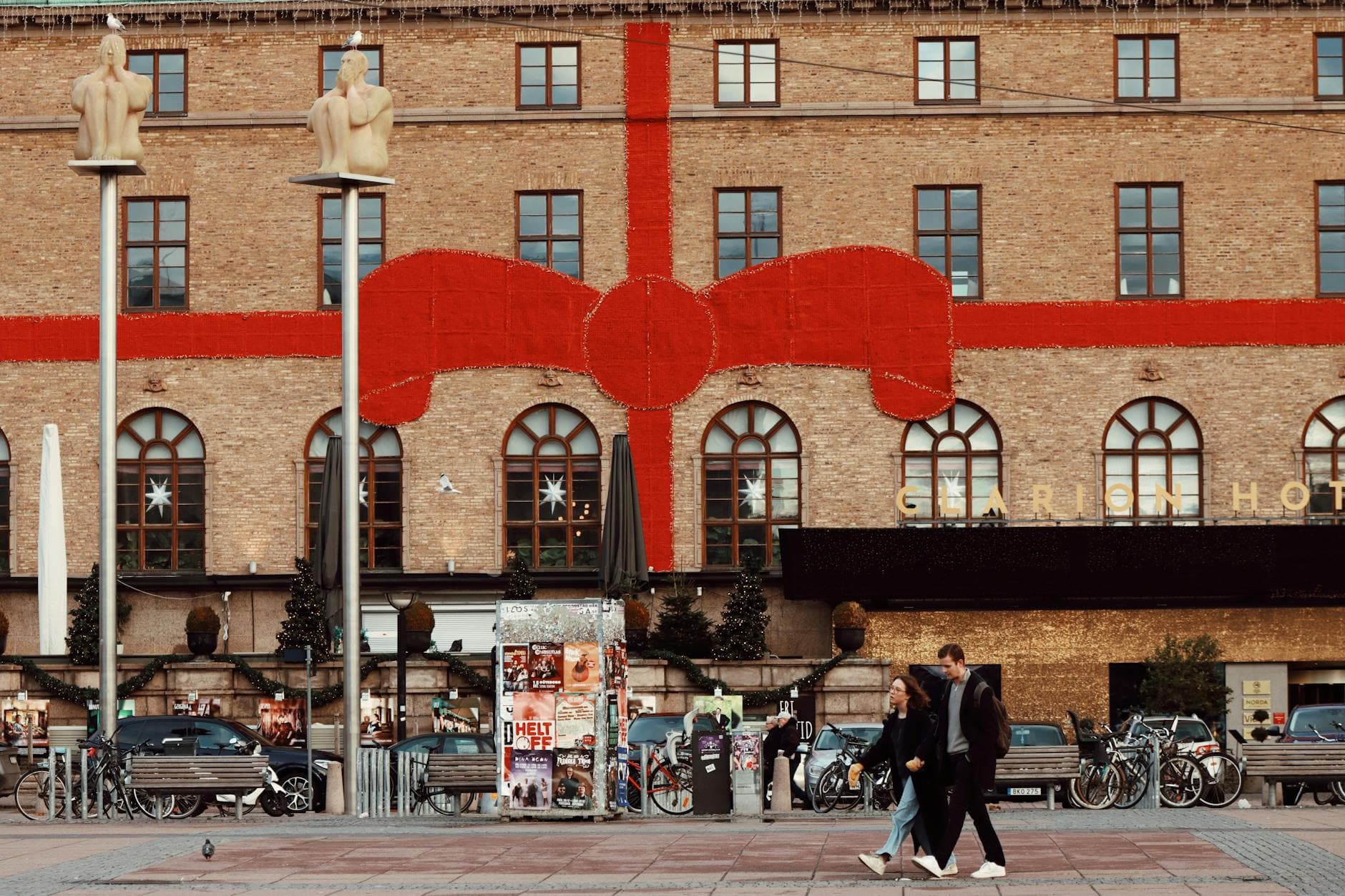 Tegelfasad inslagen i ett stort rött band medan två personer promenerar förbi på torget.