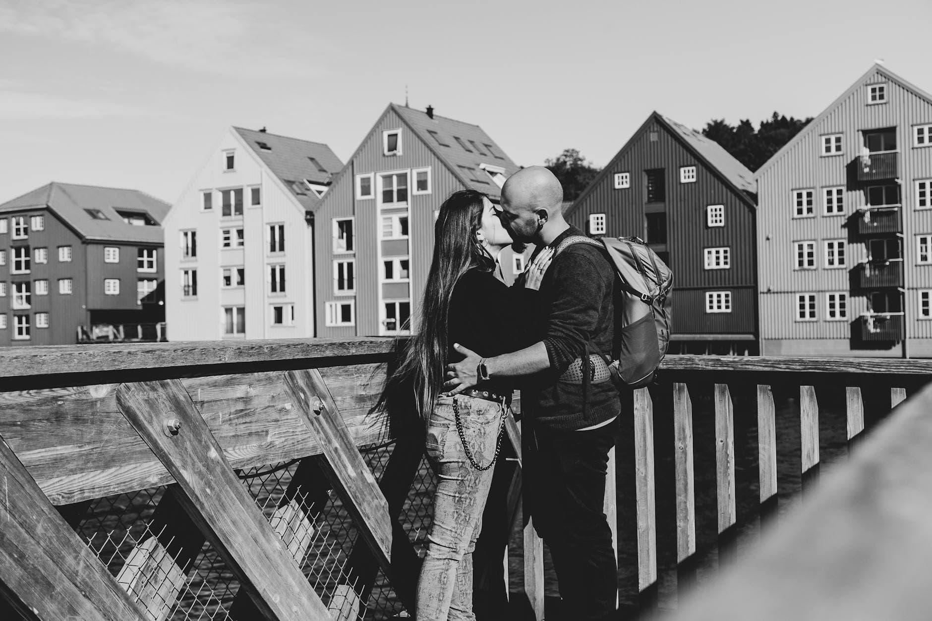 Par kysser på ei trebru foran gamle bryggehus, tatt i svart-hvitt.