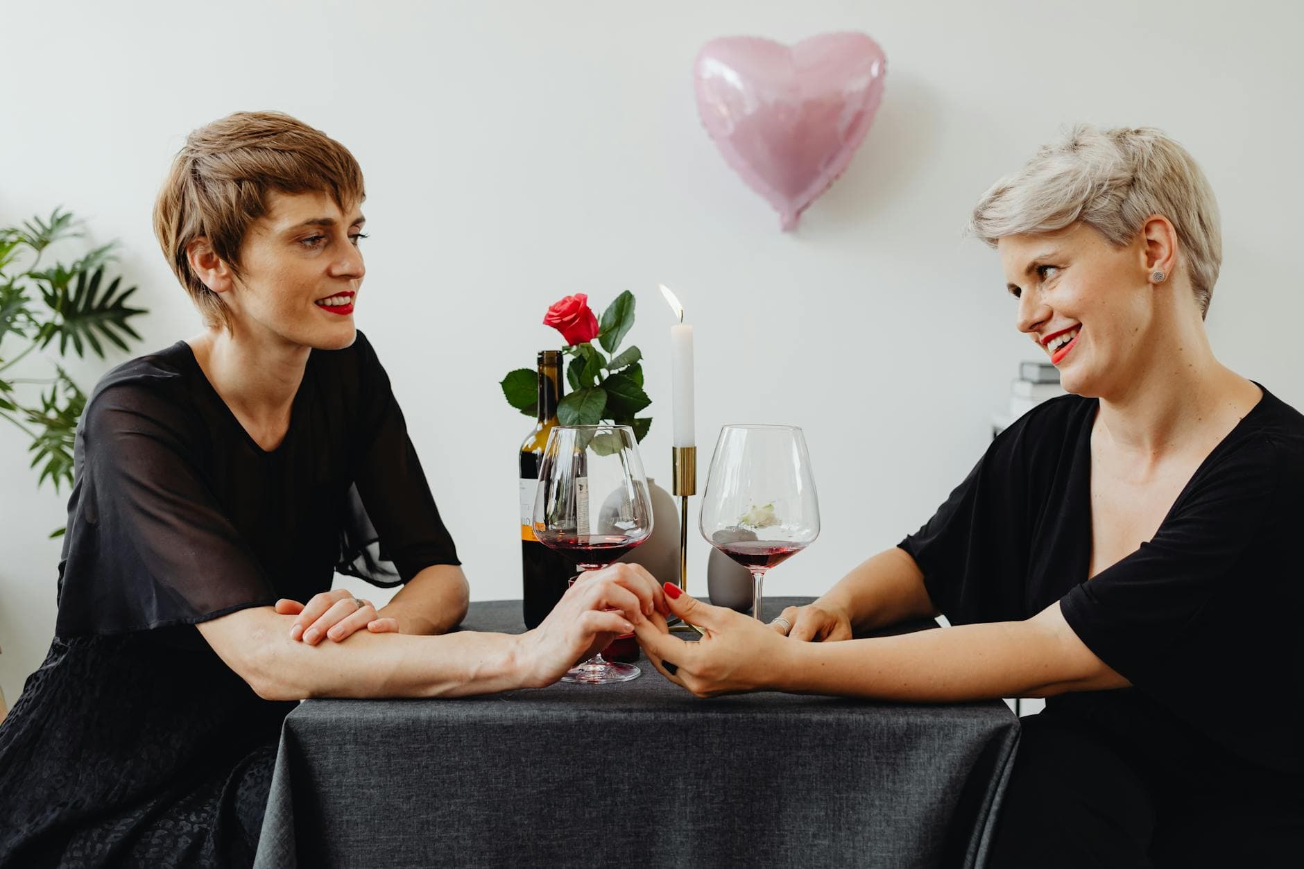 Zwei Personen in Schwarz halten am Tisch Händchen, daneben Rotwein, Rose und Herzballon.