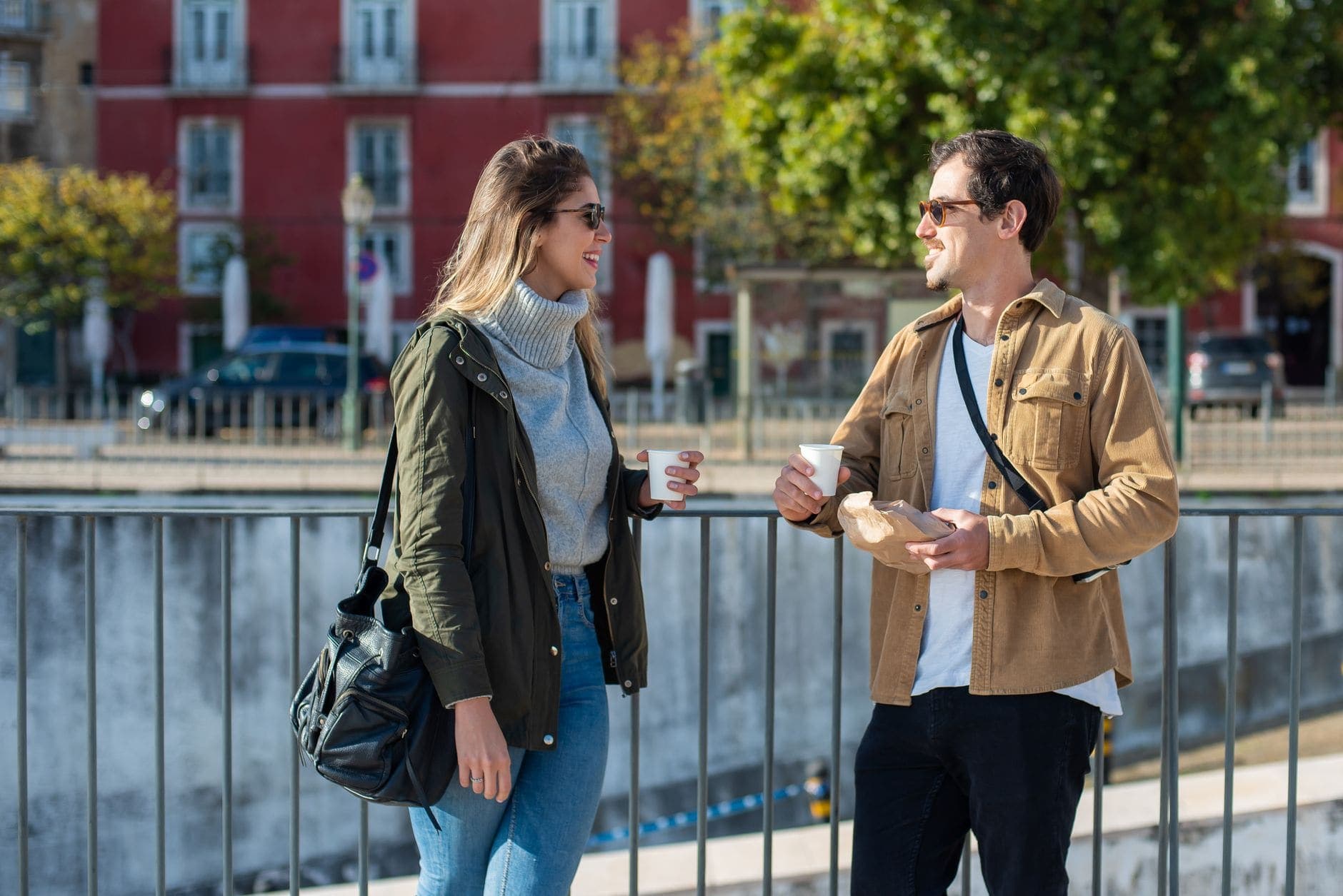 A woman and a man stand at a city railing with takeaway coffee, smiling as they talk.