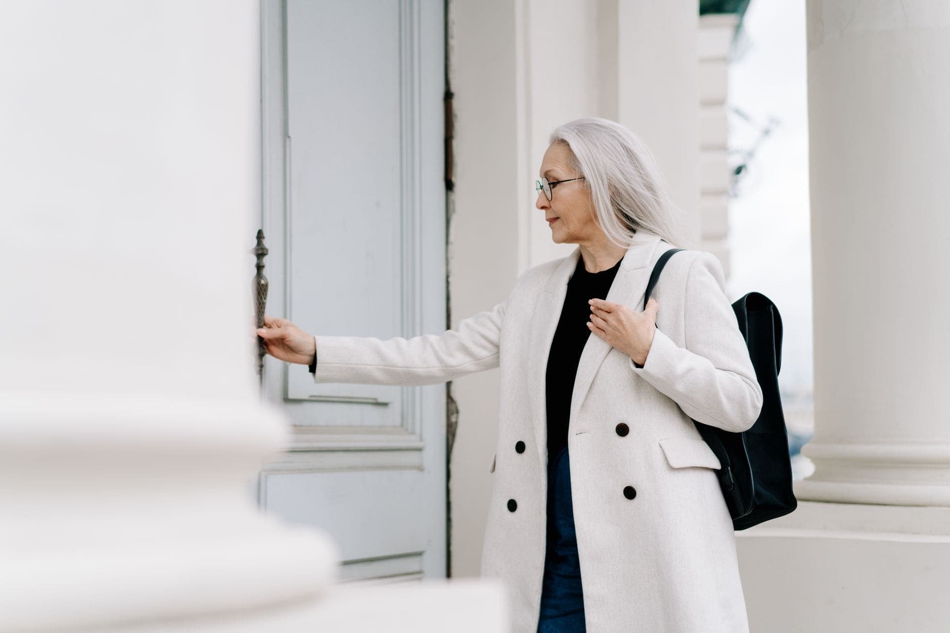Uma mulher de cabelo branco e casaco claro segura o puxador de uma porta.