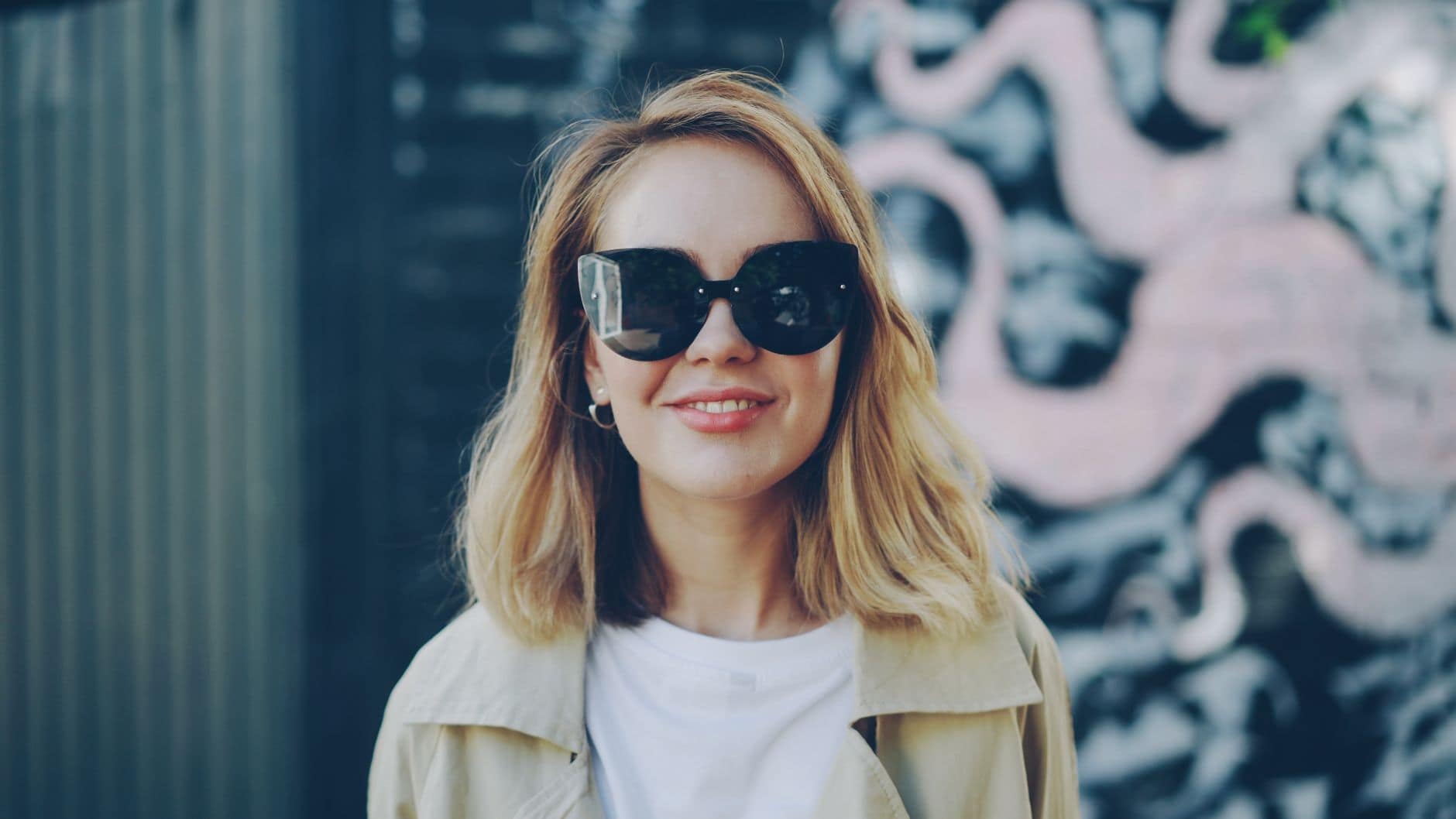 Eine Frau mit schwarzer Sonnenbrille und heller Jacke lächelt vor einer bemalten Wand.