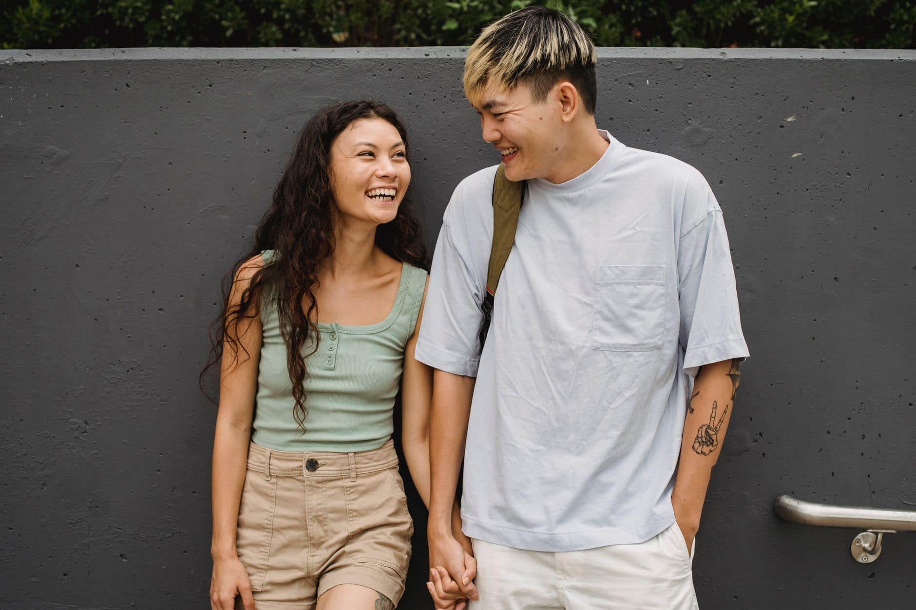 Un homme et une femme se tiennent la main et rient devant un mur gris.