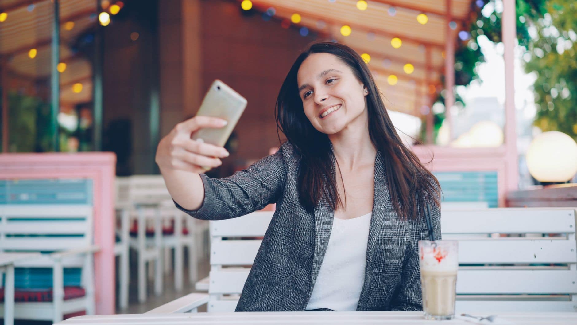 Usmiata žena v sivom károvanom saku si robí selfie pri stole v pastelovo zariadenej kaviarni
