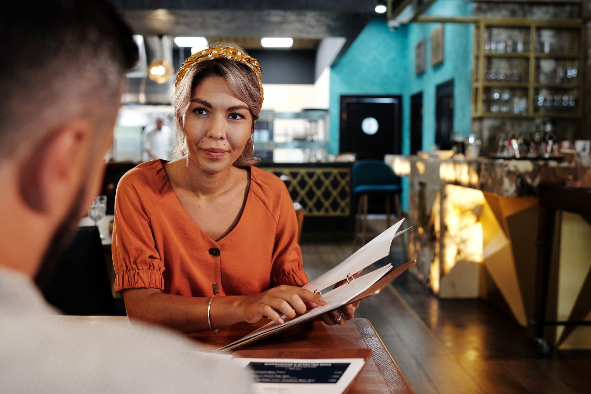 Une femme en haut orange consulte un menu en regardant un homme assis en face d'elle.