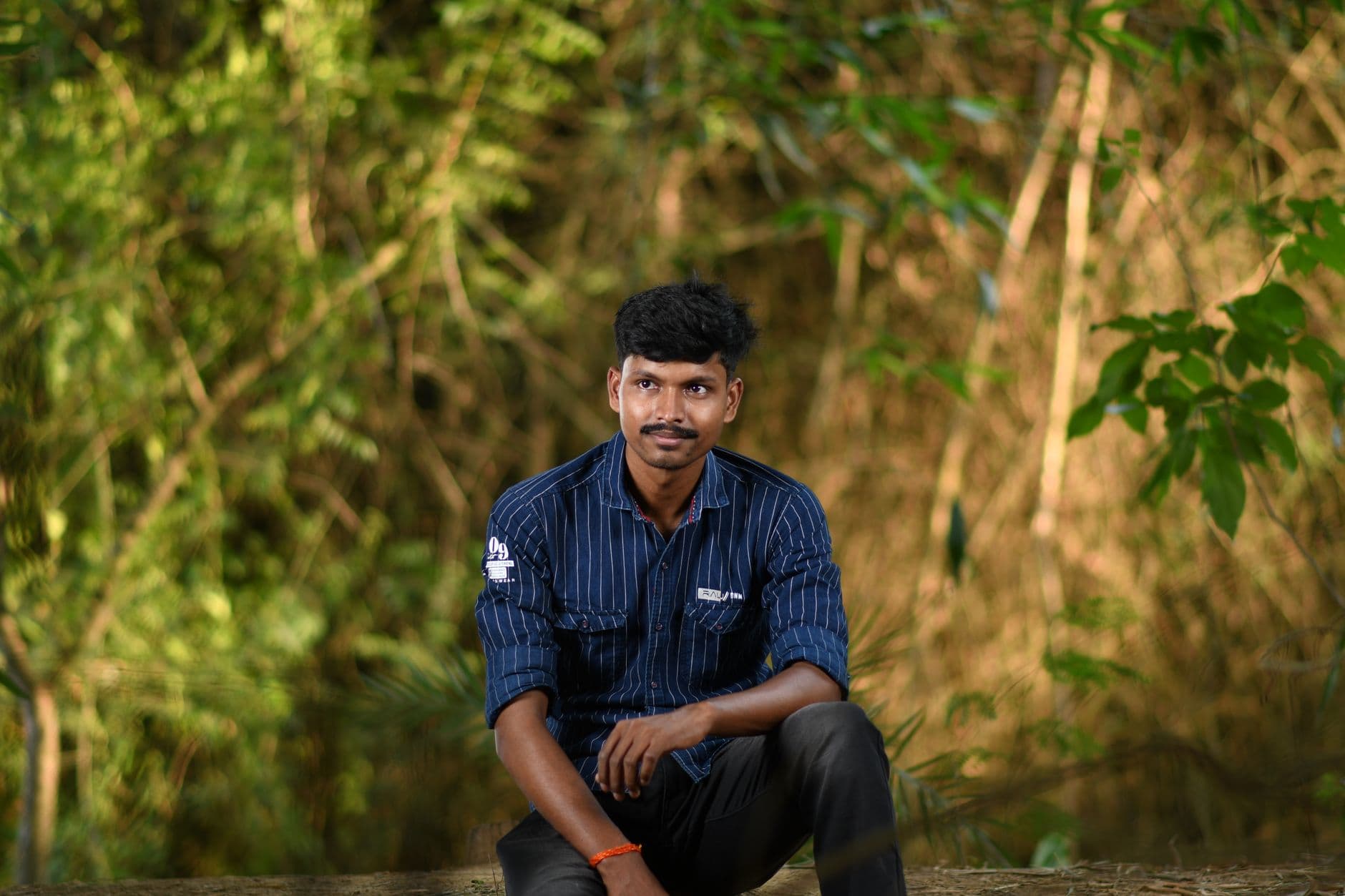 Un homme en chemise à fines rayures bleu marine est assis devant un fond de verdure.