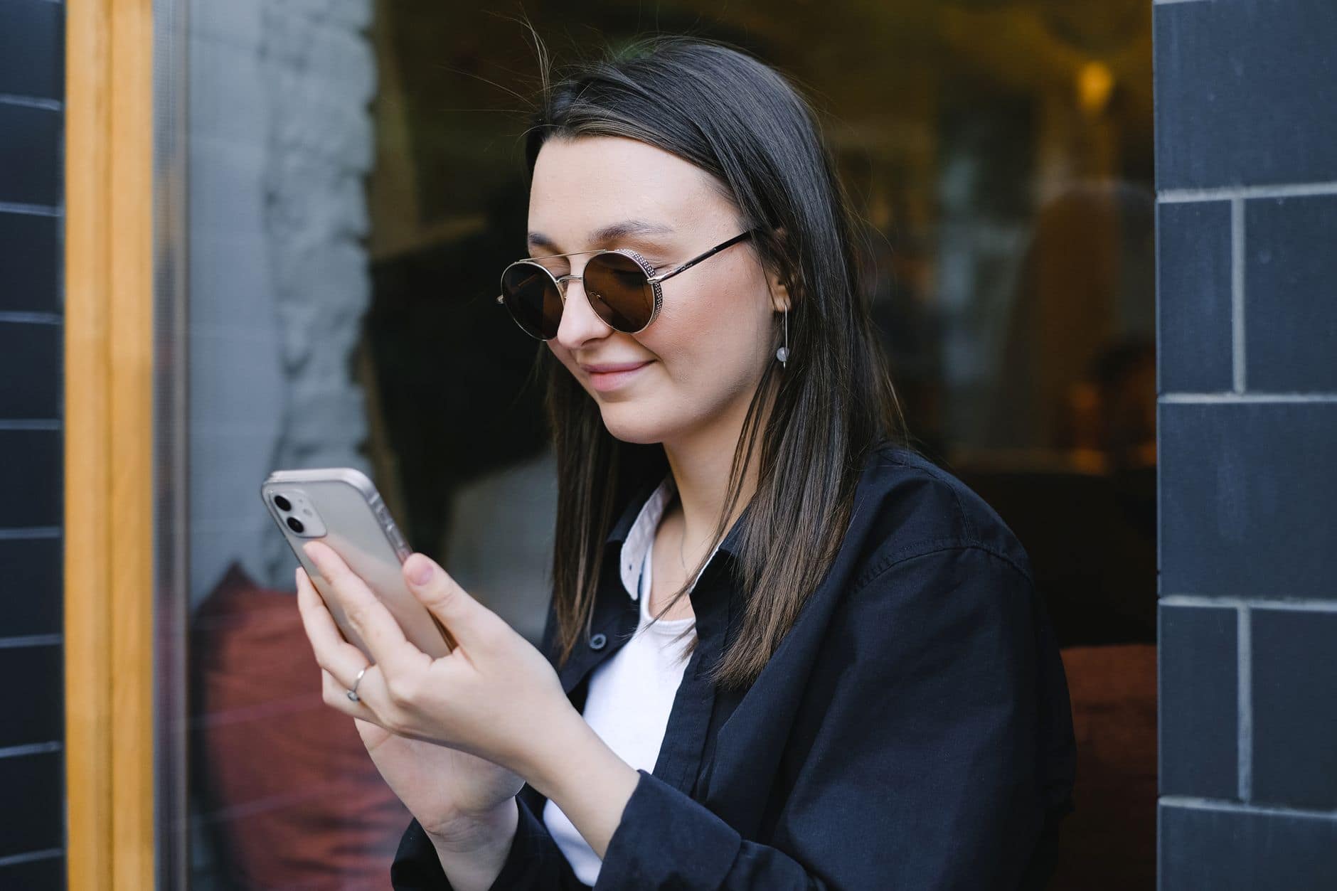 Uśmiechnięta kobieta w okrągłych okularach przeciwsłonecznych patrzy na telefon przed kawiarnią