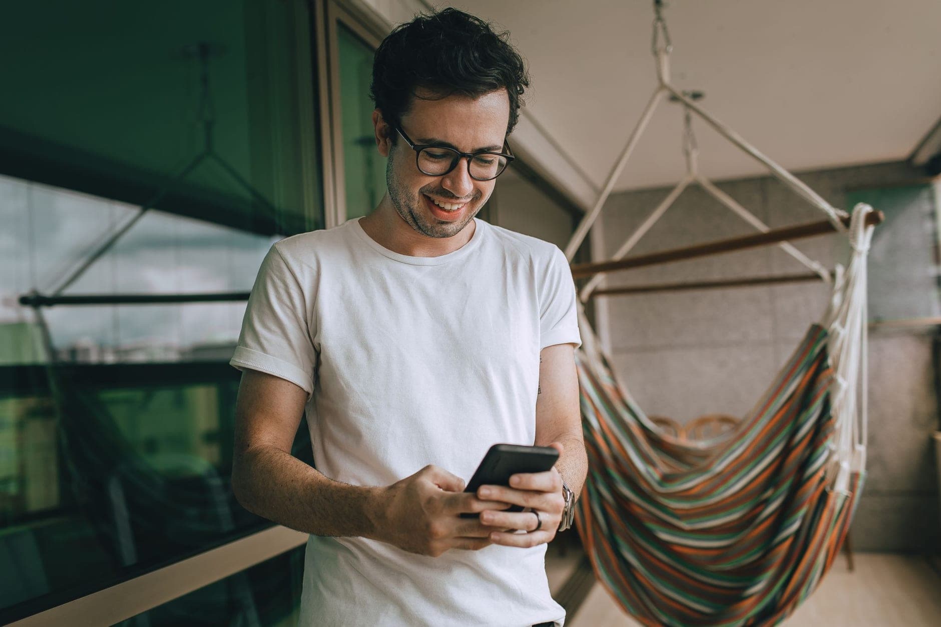 Vyras su akiniais šypsodamasis skaito telefoną terasoje šalia hamako.