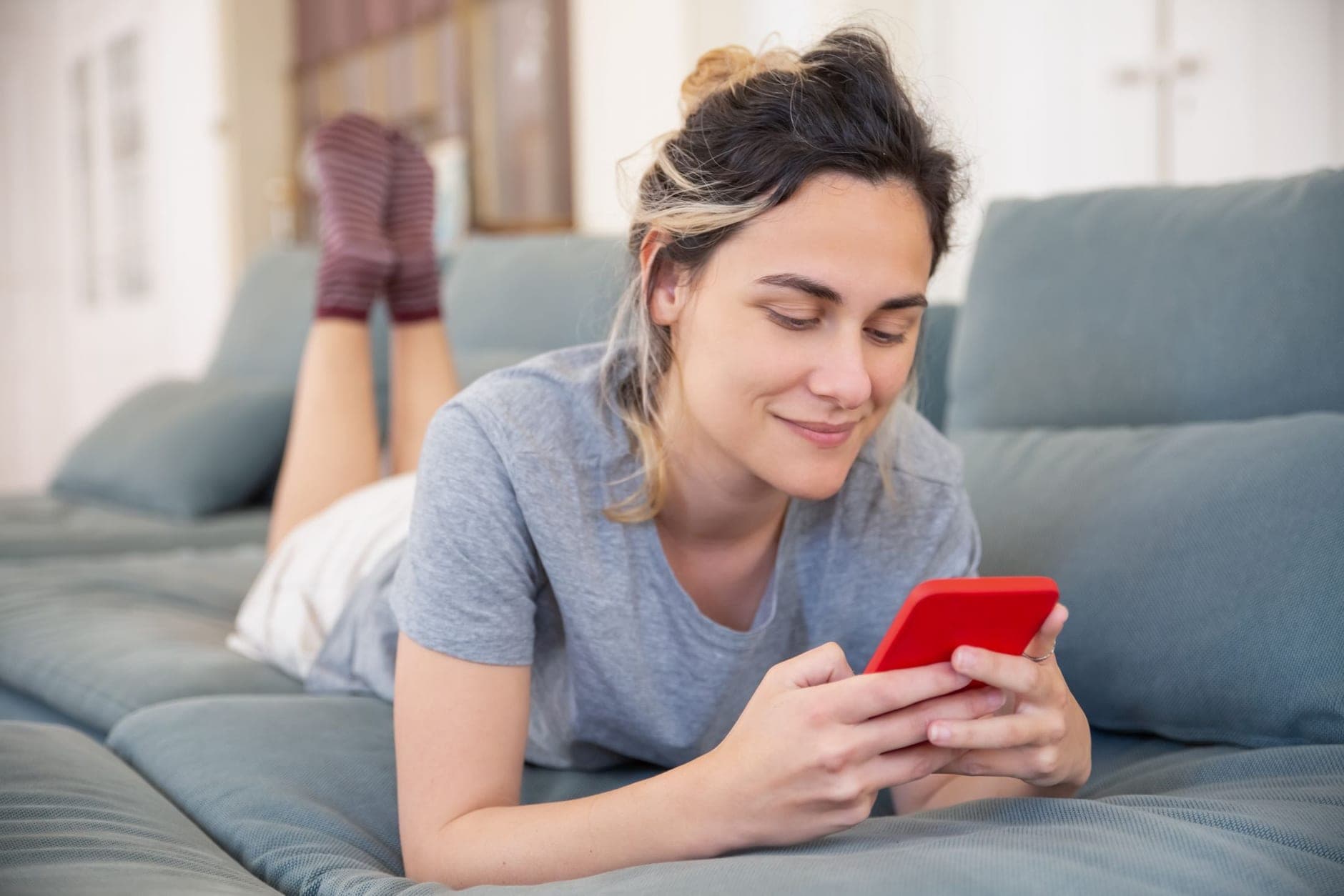 Ant sofos gulinti moteris lengvai šypsosi žiūrėdama į telefoną su raudonu dėklu.