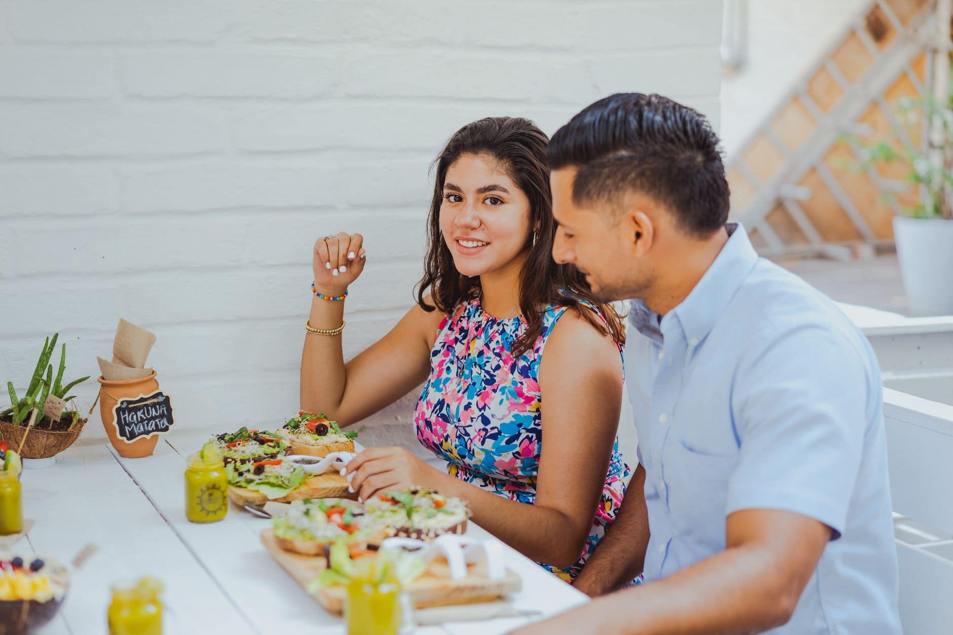 Casal almoça em uma mesa ao ar livre, a mulher sorri para a câmera ao lado dele.