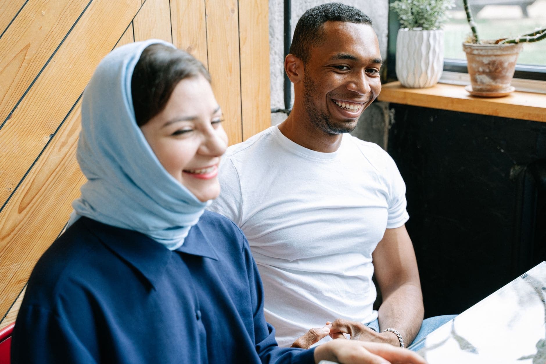 A woman in a light blue headscarf and a man in a white T-shirt smile at a bright café table by a window