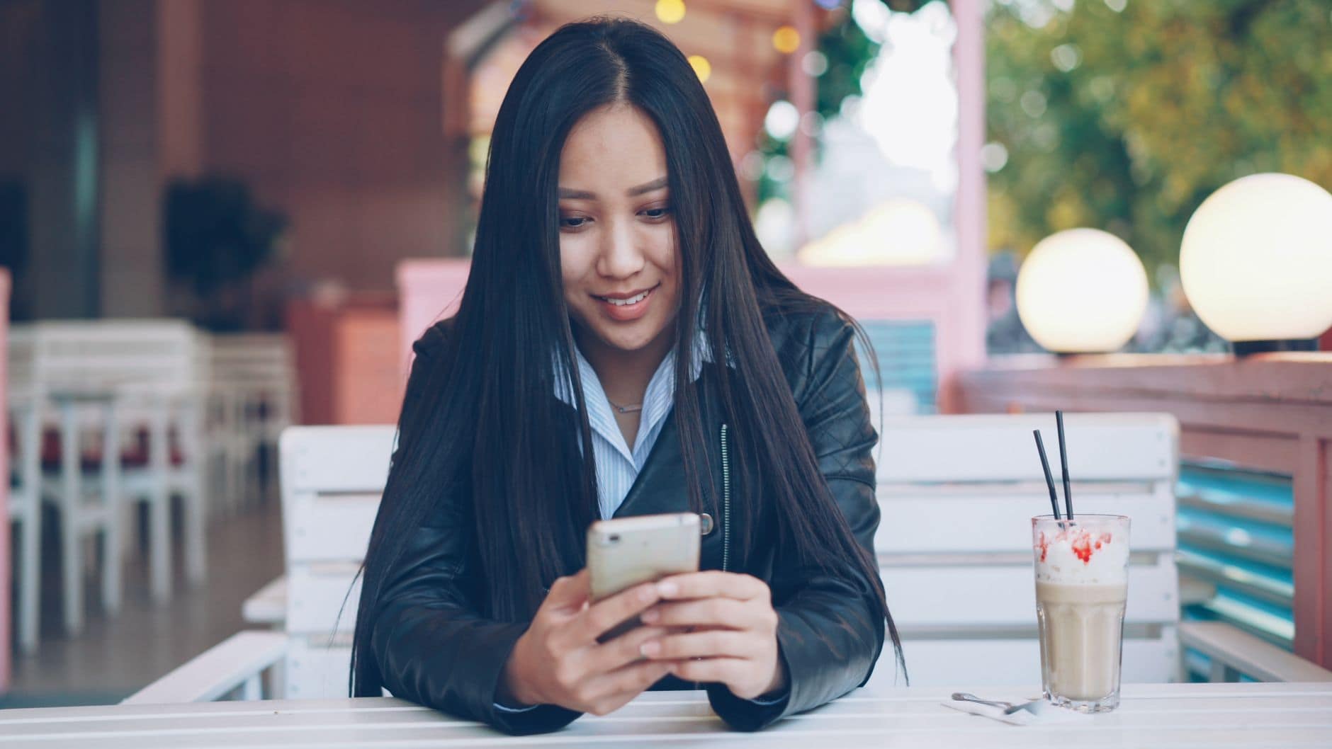 Une femme en veste noire sourit en regardant son téléphone à une table de café.