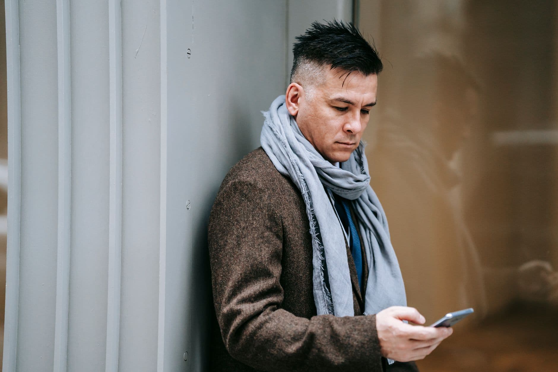 Un homme en manteau et écharpe grise consulte son téléphone dehors.