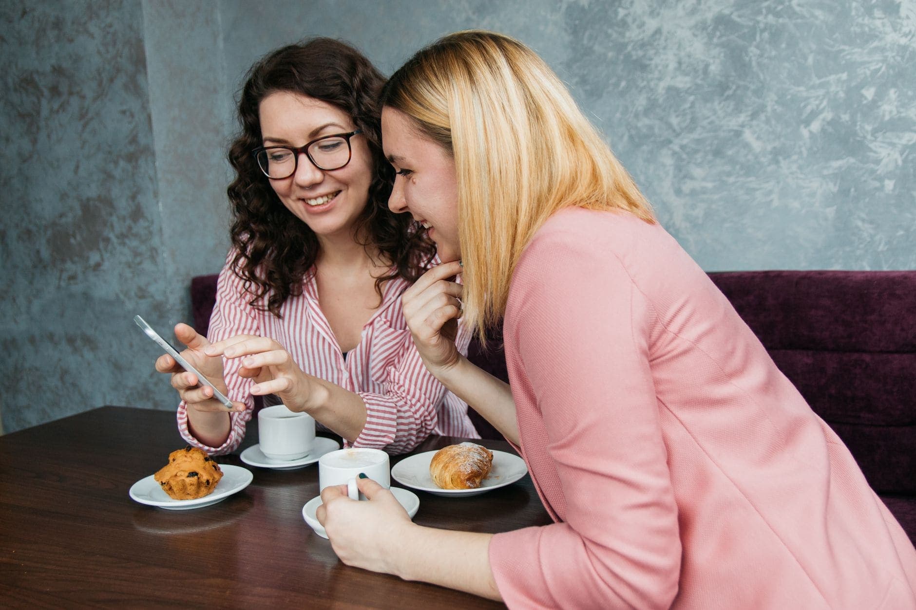 Duas mulheres olham para um telemóvel num café, com chávenas e bolos sobre a mesa.