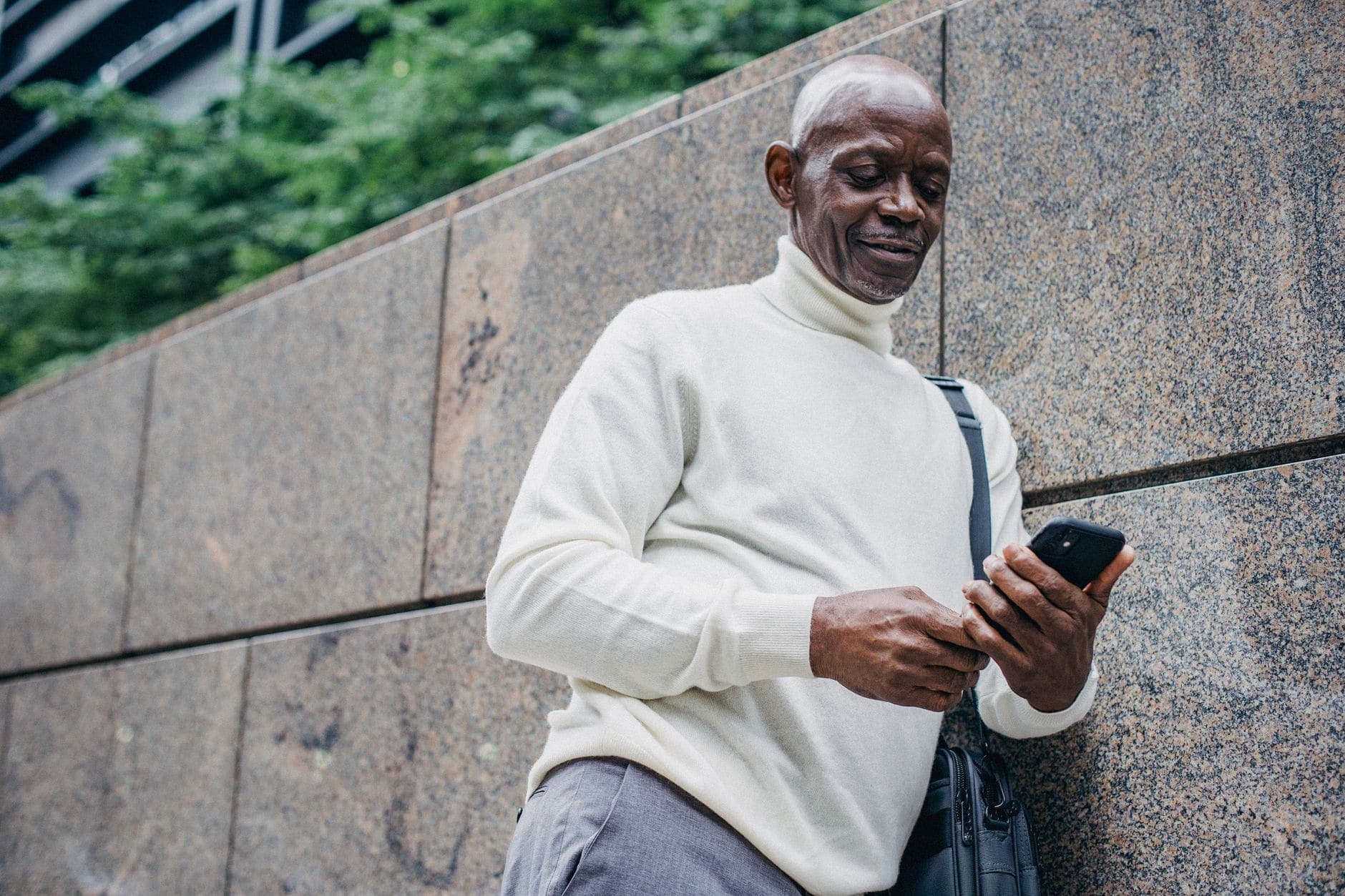 Um homem de blusa clara de gola alta olha para um celular junto a uma parede de pedra.